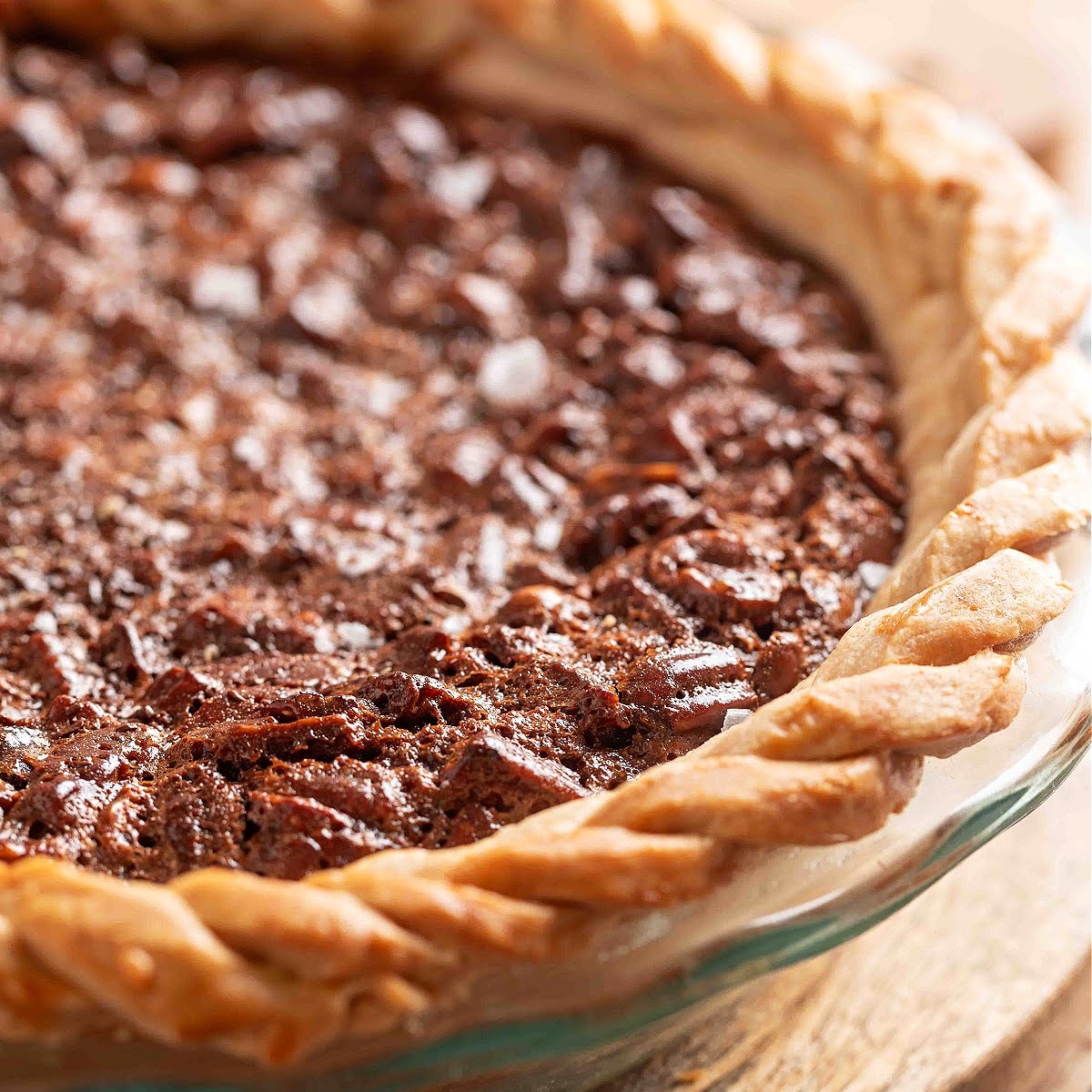 A chocolate hazelnut pecan pie in a glass pie dish with a braided crust, the top glossy and cracked with a sprinkle of flaky sea salt catching the light.