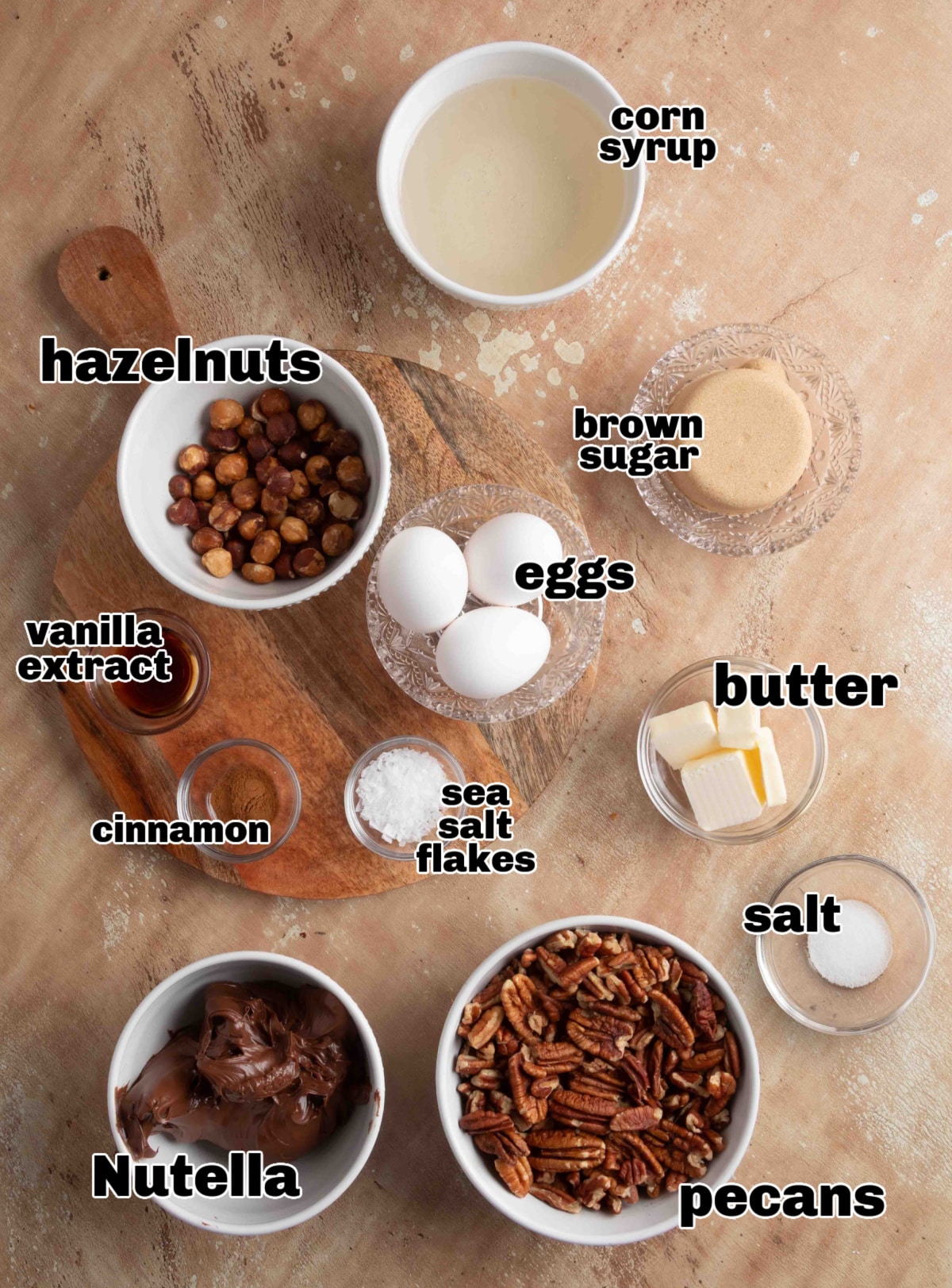 Ingredients for chocolate hazelnut pecan pie laid out on a board, including Nutella, pecans, hazelnuts, eggs, butter, brown sugar, corn syrup, cinnamon, vanilla, and sea salt.