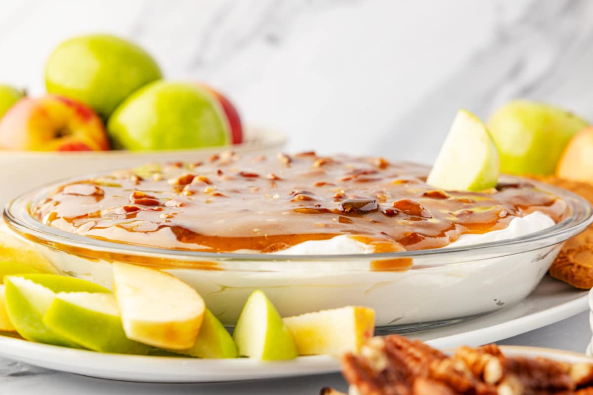 Pecan topping layered over cream cheese dip with apple slices Pecan pie dip in a glass pie dish with a cold cream cheese base topped with warm caramel pecans, surrounded by sliced green apples for dipping by Noble Pig.