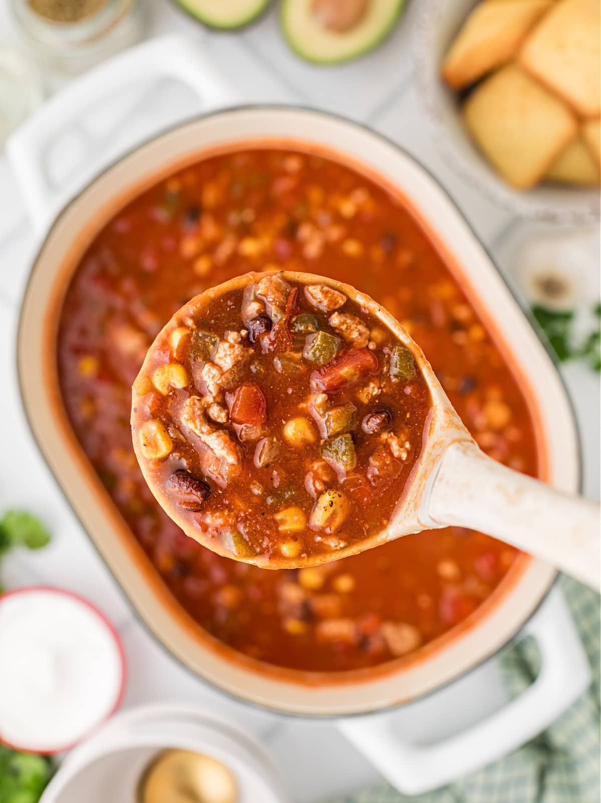 A ladle of ground chicken chili with black beans, corn, and tomatoes held over a Dutch oven filled with chili for Noble Pig.