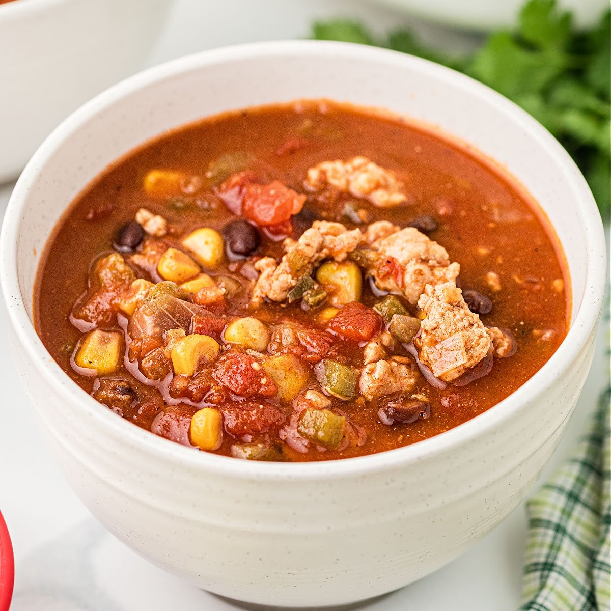 A white bowl of hearty ground chicken chili with corn, black beans, and tomatoes in a rich red broth.