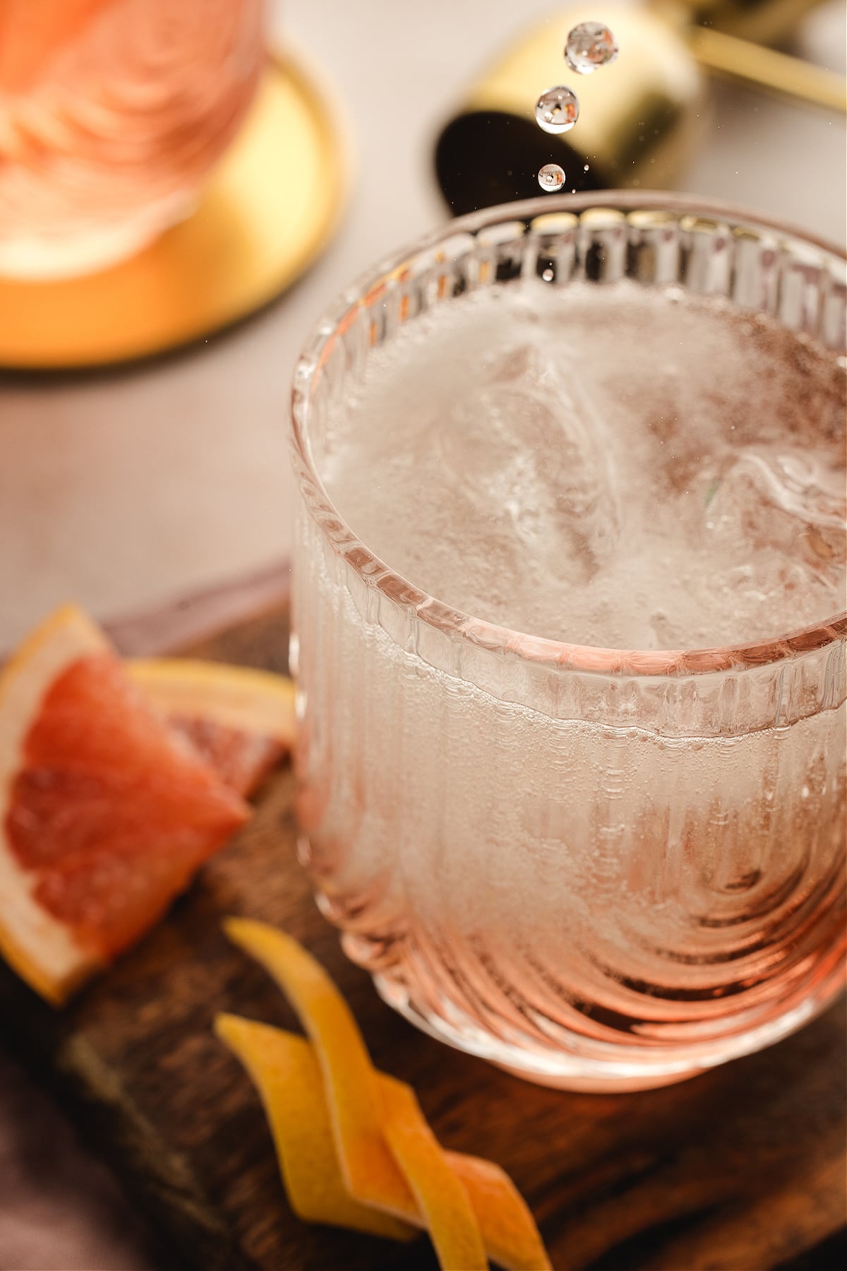 A chilled Lillet Rosé spritz catching falling droplets, the pale pink drink bubbling over ice with grapefruit slices and twists scattered on the wooden board below.
