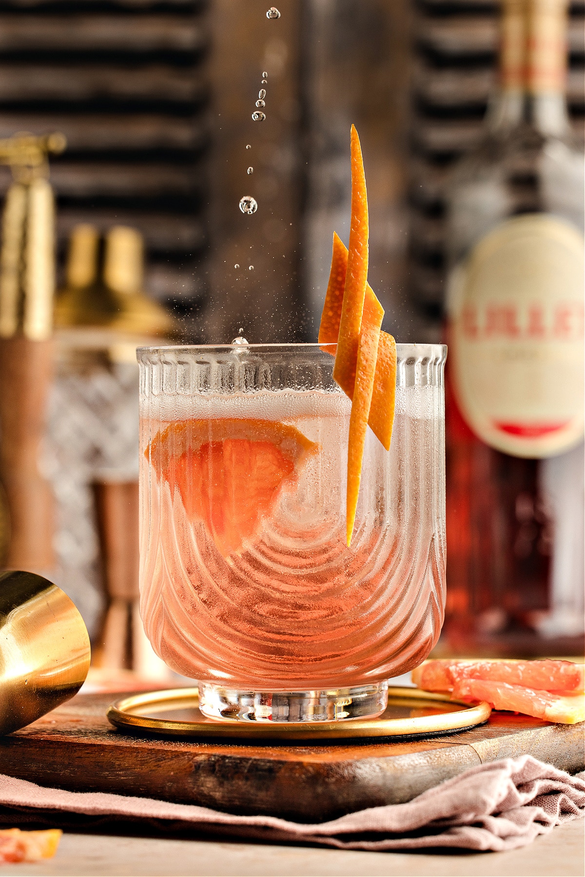 A chilled Lillet Rosé spritz in a textured glass with grapefruit garnish, catching falling droplets as the pale pink drink glows against a blurred bar background.