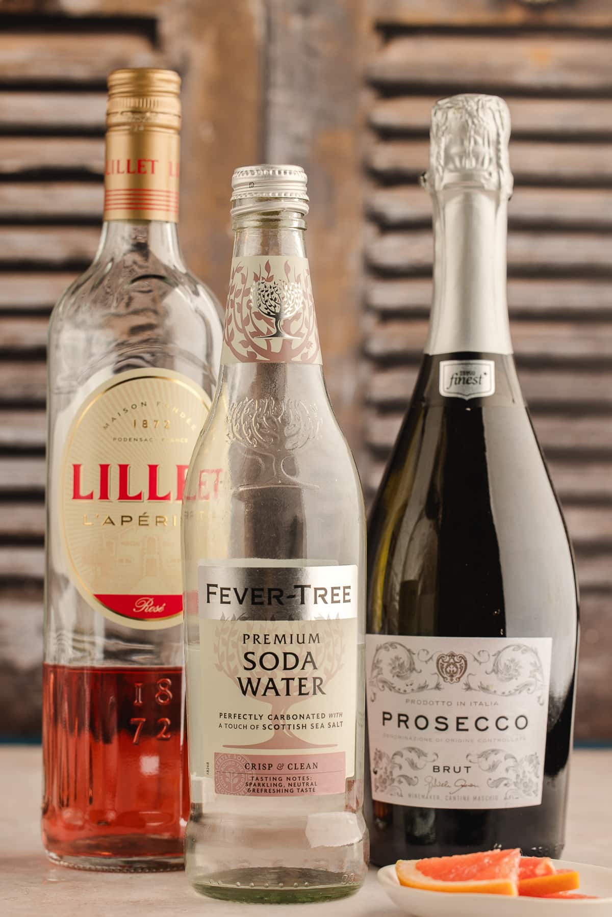 The three ingredients for a Lillet Rosé spritz—Lillet Rosé, club soda, and a bottle of brut prosecco—standing together with fresh grapefruit slices nearby.