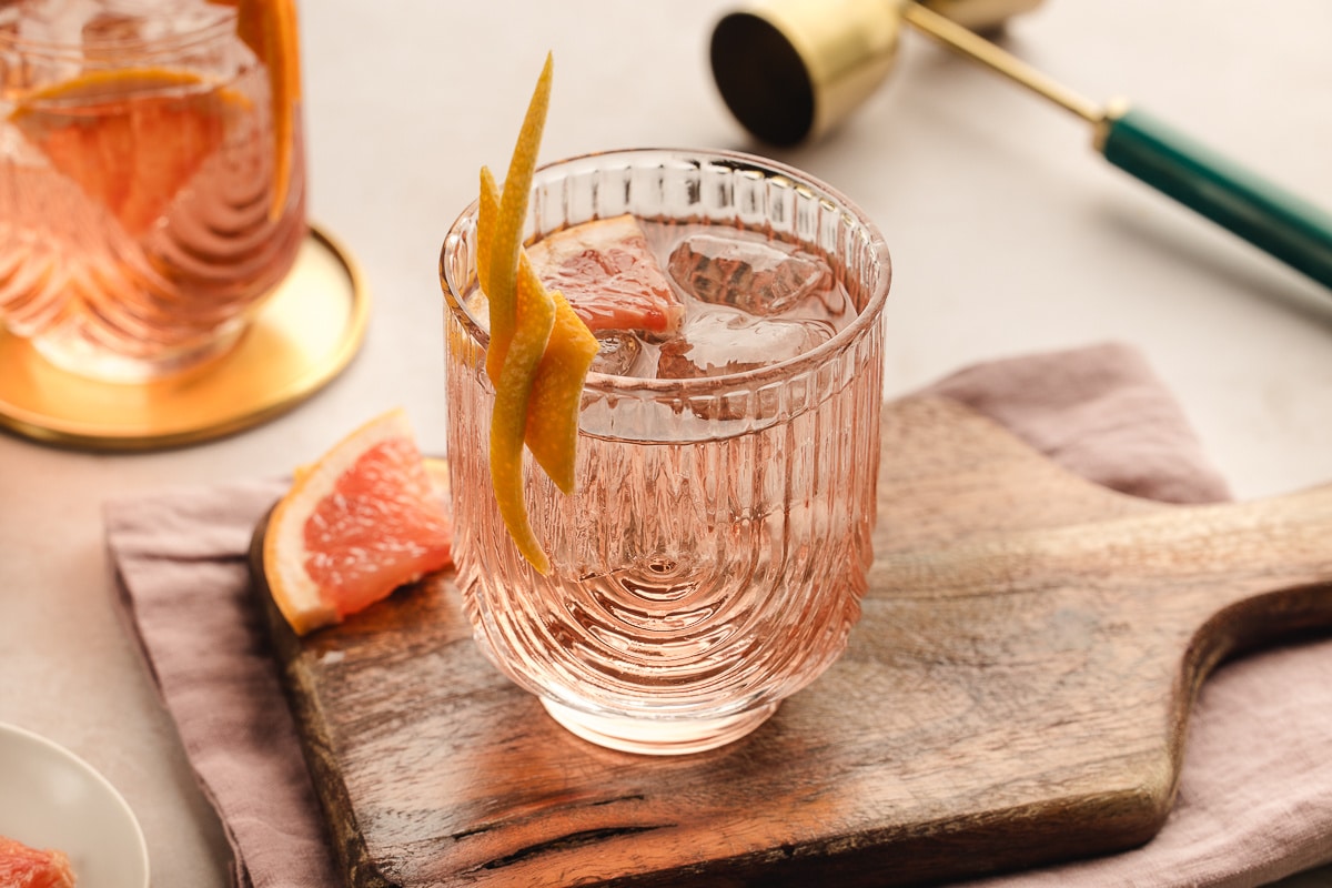 A pale pink Lillet Rosé spritz served over ice in a textured glass, garnished with a long grapefruit twist and set on a wooden board with fresh grapefruit nearby. 