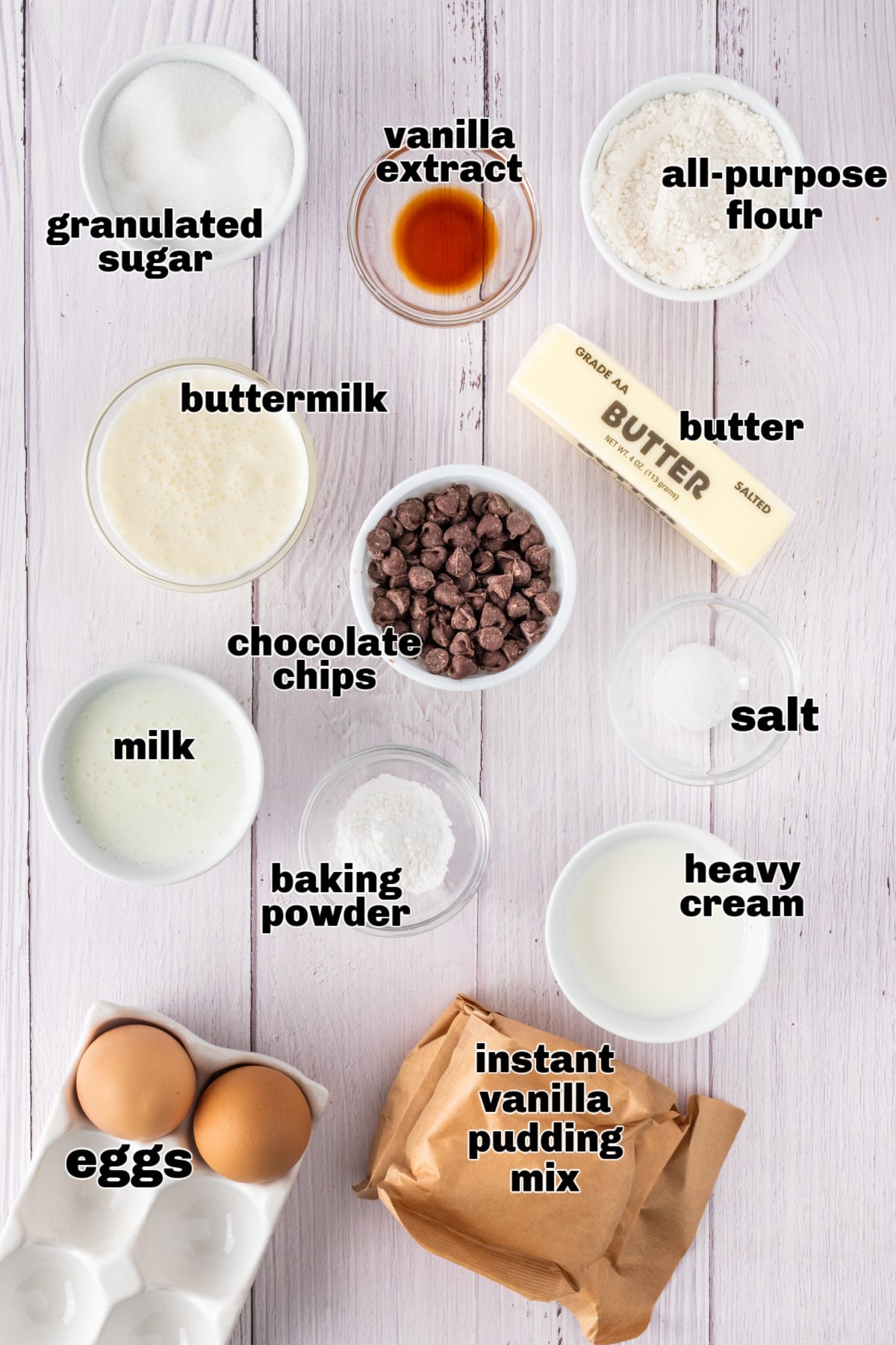 Boston Cream Pie Cupcake Ingredients Overhead view of labeled ingredients for Boston cream pie cupcakes, including flour, sugar, butter, eggs, instant vanilla pudding mix, chocolate chips, milk, and buttermilk arranged on a light wooden surface