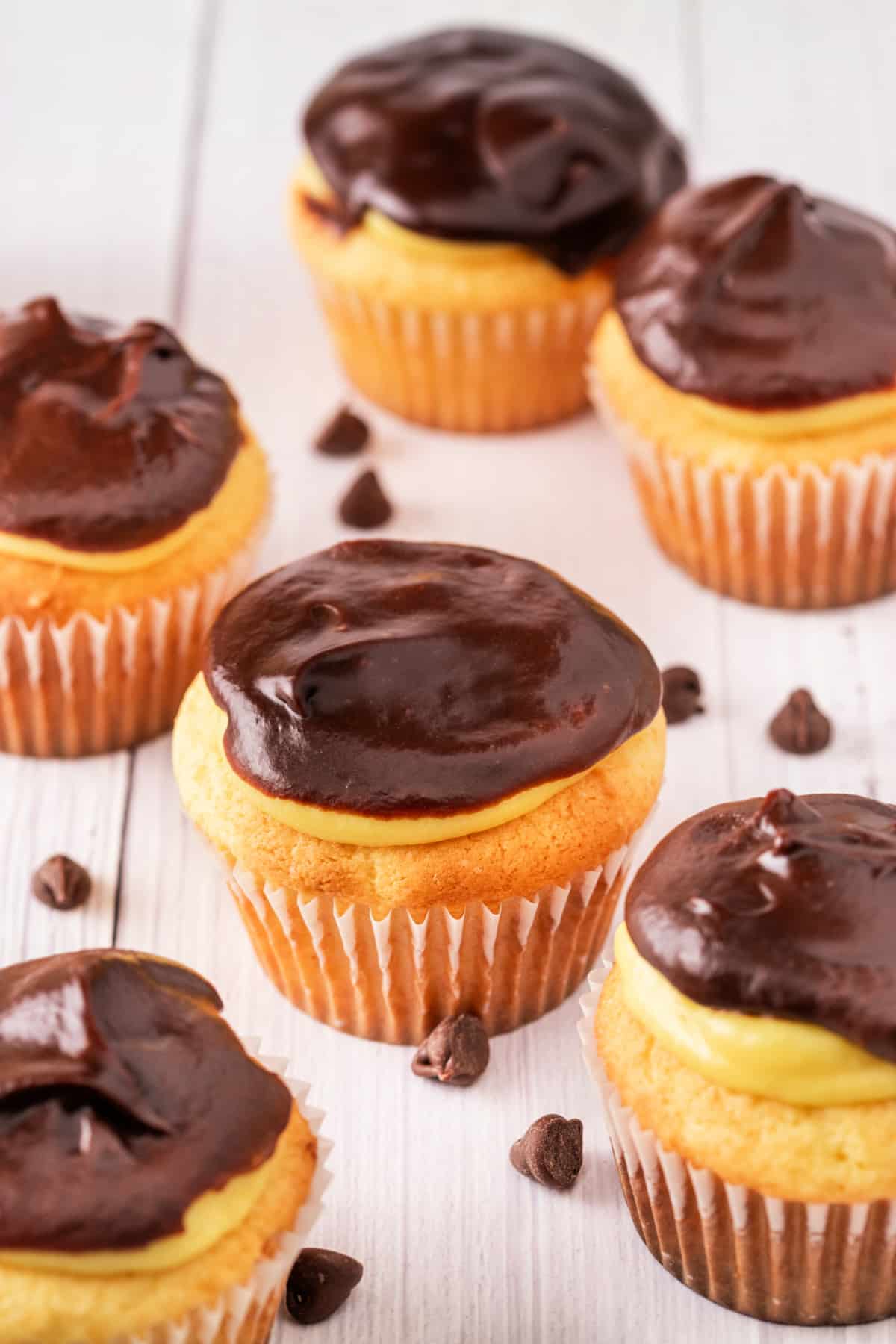 Easy Boston Cream Pie Cupcakes with Ganache Group of cupcakes with vanilla pudding filling and smooth chocolate ganache topping, arranged on a white wood surface with chocolate chips scattered around