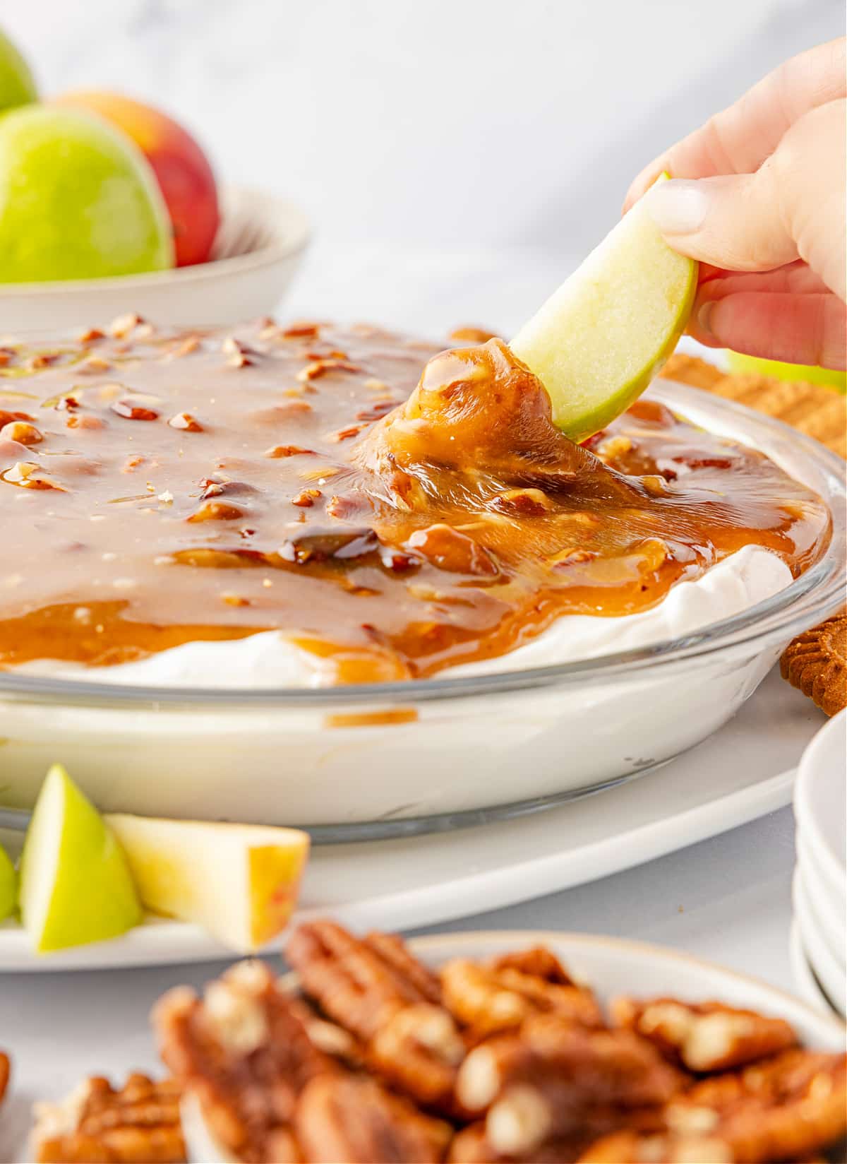 Apple slice dipped into caramel pecan topping over cream cheese layer Pecan pie dip with a cold cream cheese base topped with warm caramel pecans as a green apple slice is dipped into the topping, showing the gooey texture by Noble Pig.
