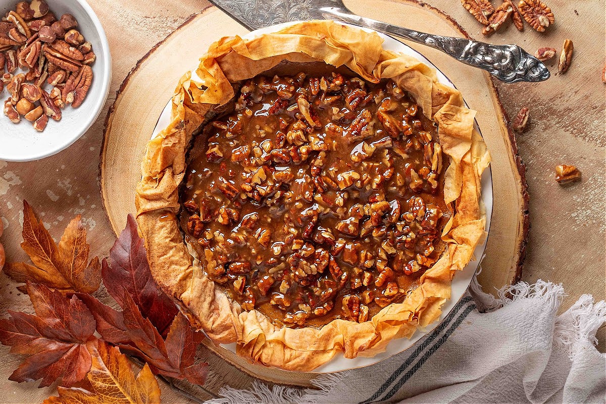 Overhead view of a pumpkin praline pie with a ruffled phyllo crust, filled with glossy brown-sugar pecan praline topping, shown on a wood slice with scattered fall leaves, pecans, and a bowl of chopped pecans nearby.