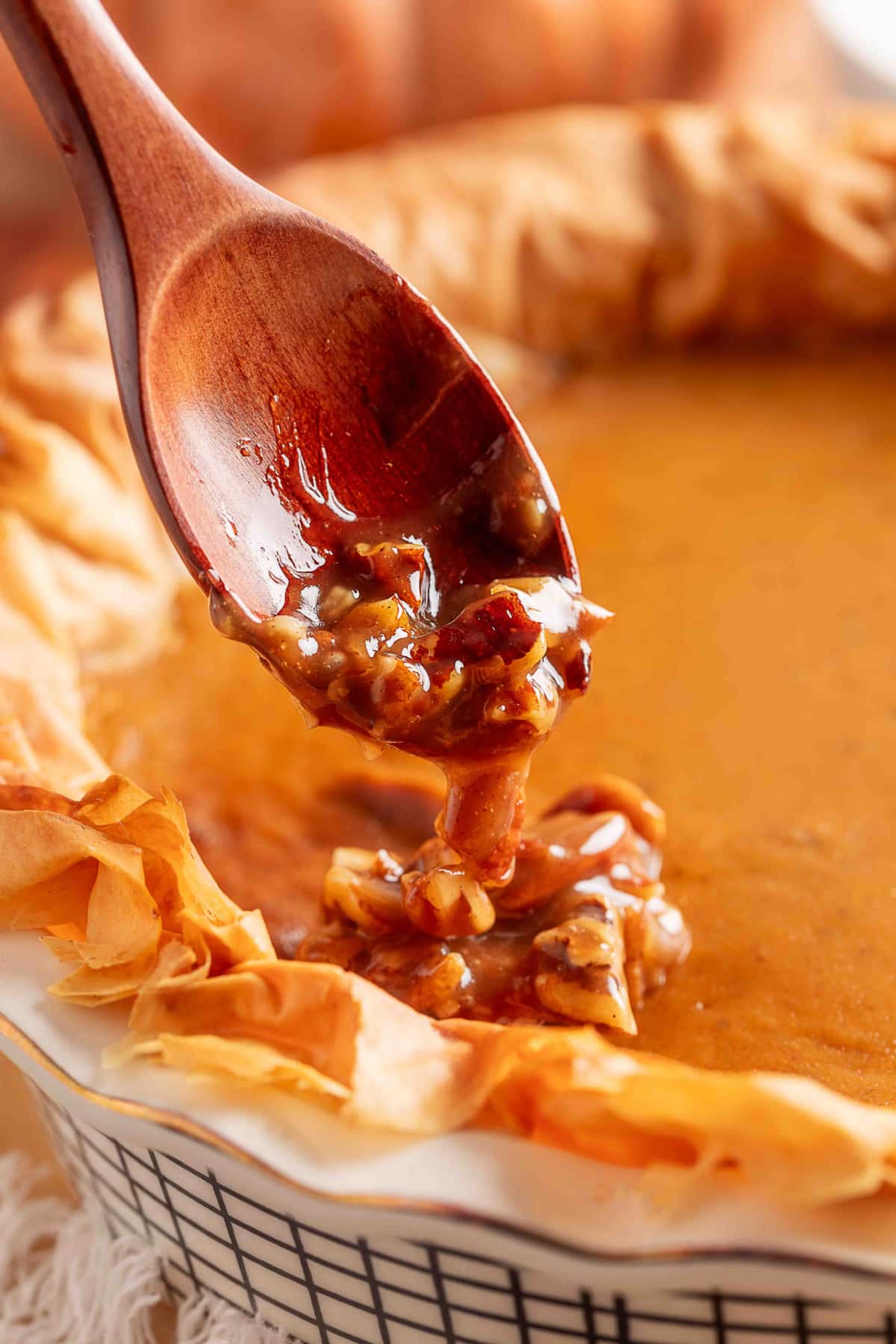 Close-up of warm pecan praline topping being spooned over a baked pumpkin pie with a ruffled phyllo crust, showing the glossy caramel coating and chopped pecans dripping from a wooden spoon onto the smooth pumpkin surface.
