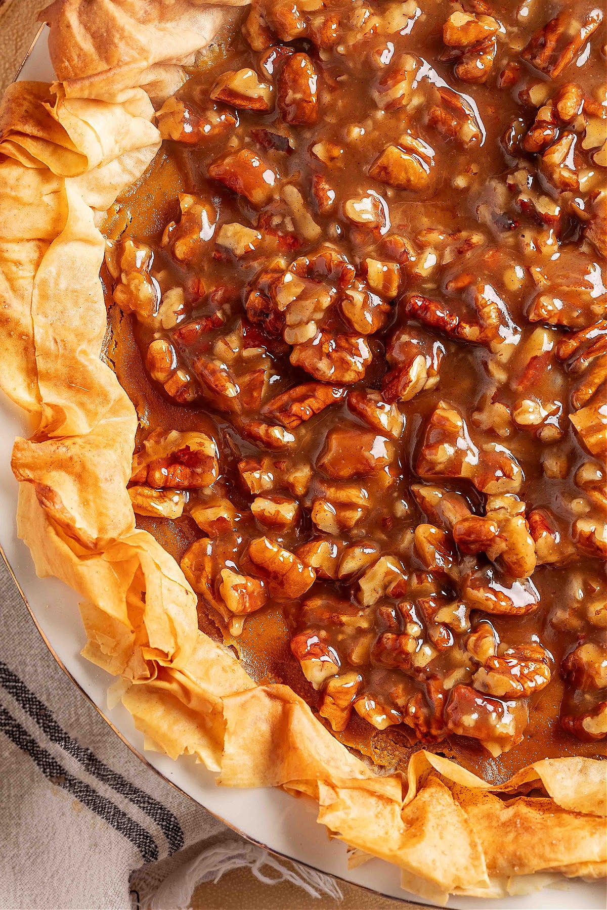 A close-up of a pumpkin praline pie with a crackling phyllo crust and glossy pecan praline topping, showing the ruffled, golden phyllo edges and the thick, caramel-like pecan layer settling into the pumpkin custard beneath.