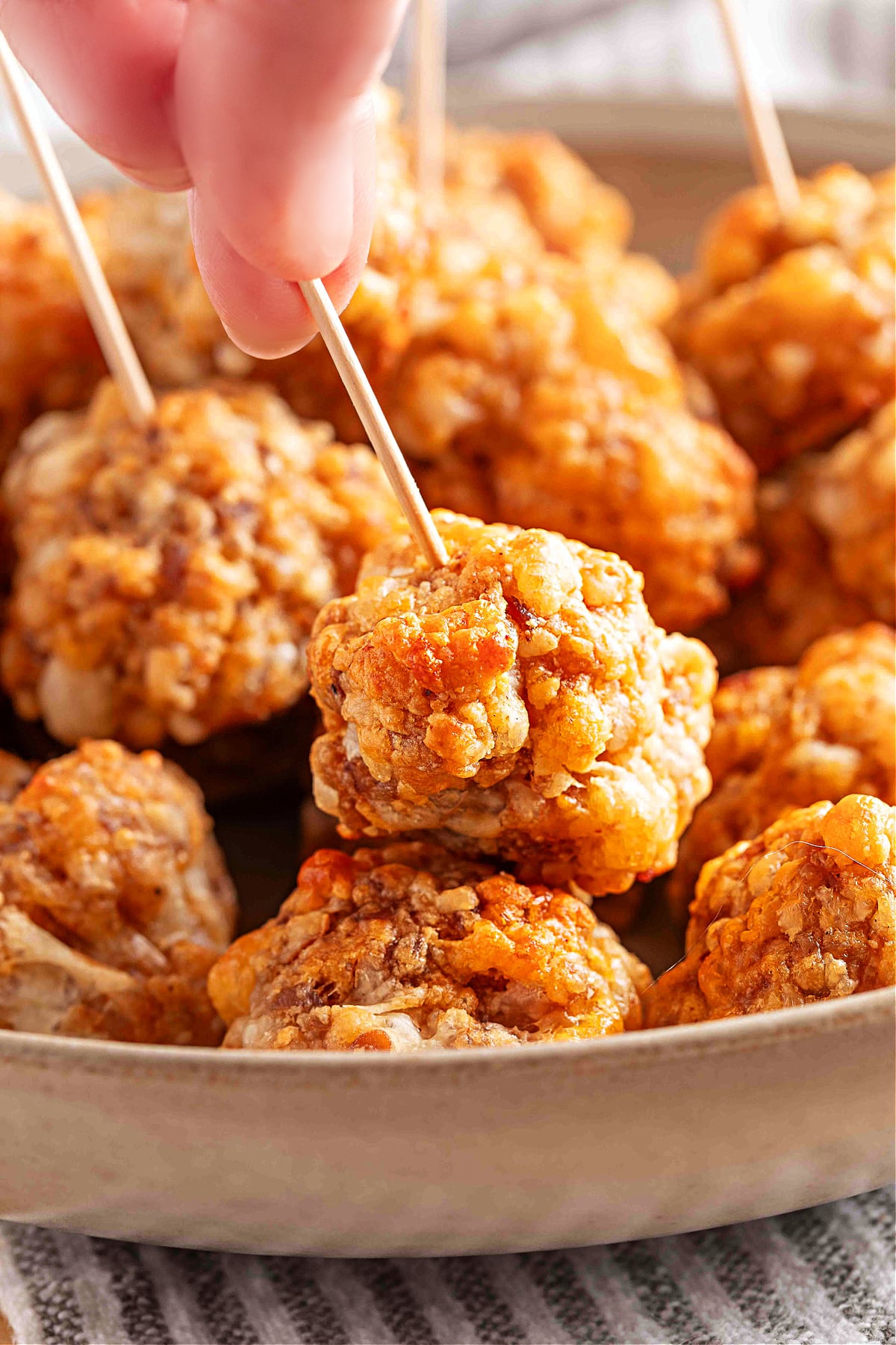 A toothpick lifting a golden, crispy sausage ball from a serving bowl. A hand picks up a smoked cheddar sausage ball with a toothpick, showing its crisp, golden surface and tender baked texture, by Noble Pig.
