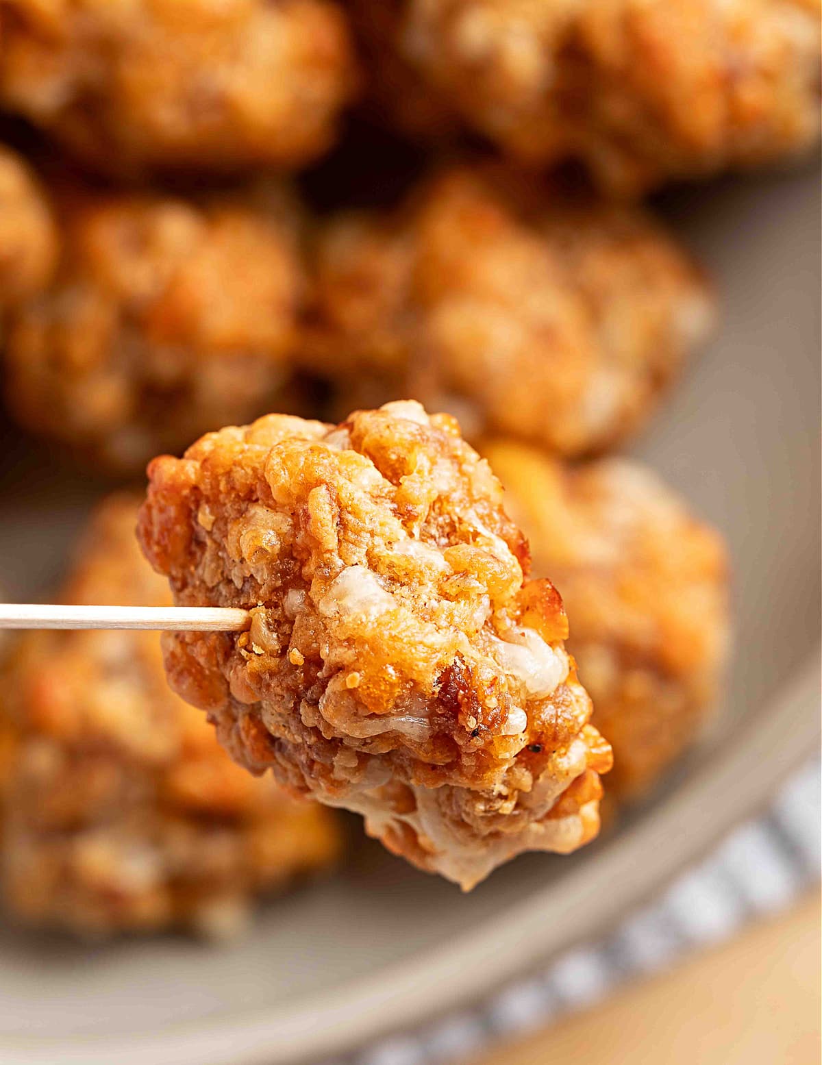 A sausage ball held on a toothpick over a bowl of freshly baked sausage balls. A single sausage ball held on a toothpick, showing its crisp, golden exterior and melted smoked cheddar against a blurred bowl of more sausage balls.