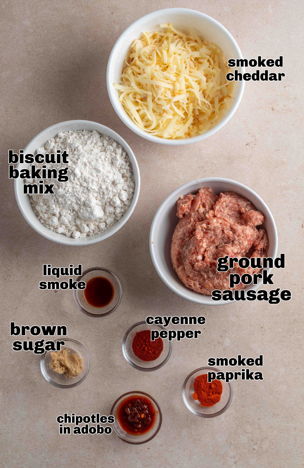Ingredients for smoked cheddar sausage balls laid out in small bowls. Ingredients for smoked cheddar sausage balls arranged on a countertop, including biscuit baking mix, ground pork sausage, shredded smoked cheddar, chipotle in adobo, brown sugar, smoked paprika, cayenne, and liquid smoke.