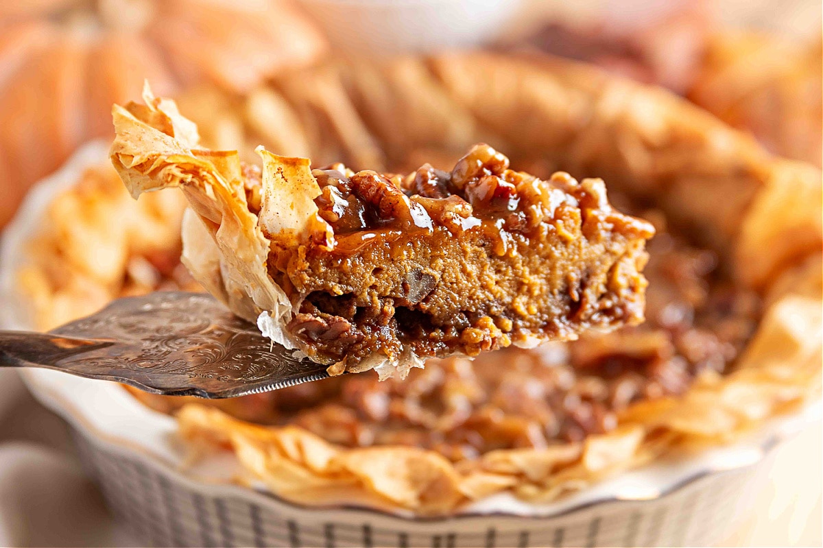 A close-up slice of pumpkin praline pie being lifted from the dish, showing the crisp, flaky phyllo crust, the pecan-studded brown-sugar base, the smooth pumpkin custard center, and the glossy praline topping dripping over the edges.