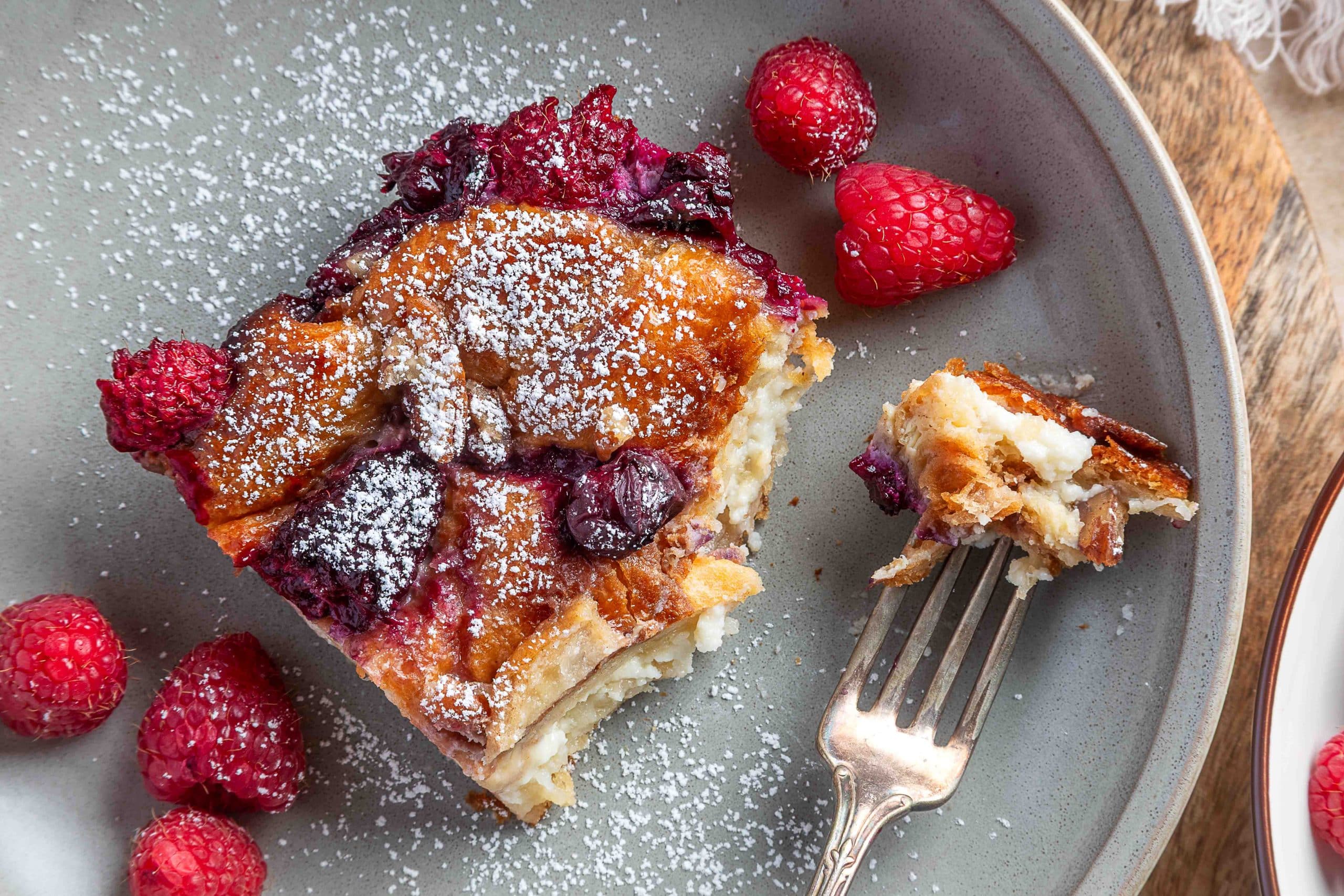 A spoon scoops into the baked croissant French toast casserole in the dish, showing the custardy interior with berries and pecans on top.