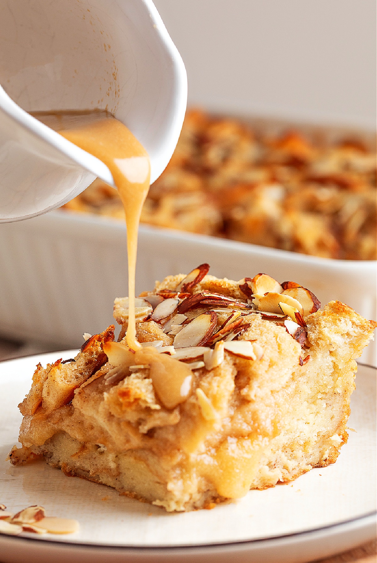 Warm brown sugar glaze being poured over a slice of almond-topped bread pudding, settling into the soft layers while the rest of the pan sits blurred in the background.
