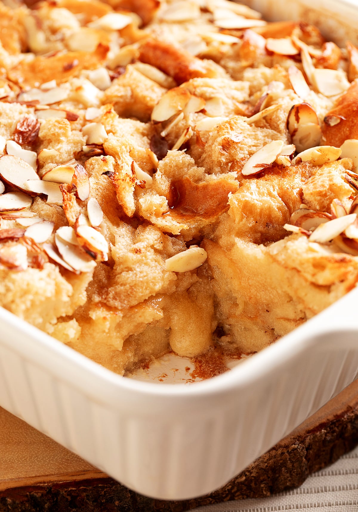 A close-up look at the baked almond cream bread pudding, showing the toasted sliced almonds on top and the soft, custardy interior where a piece has been scooped out.
