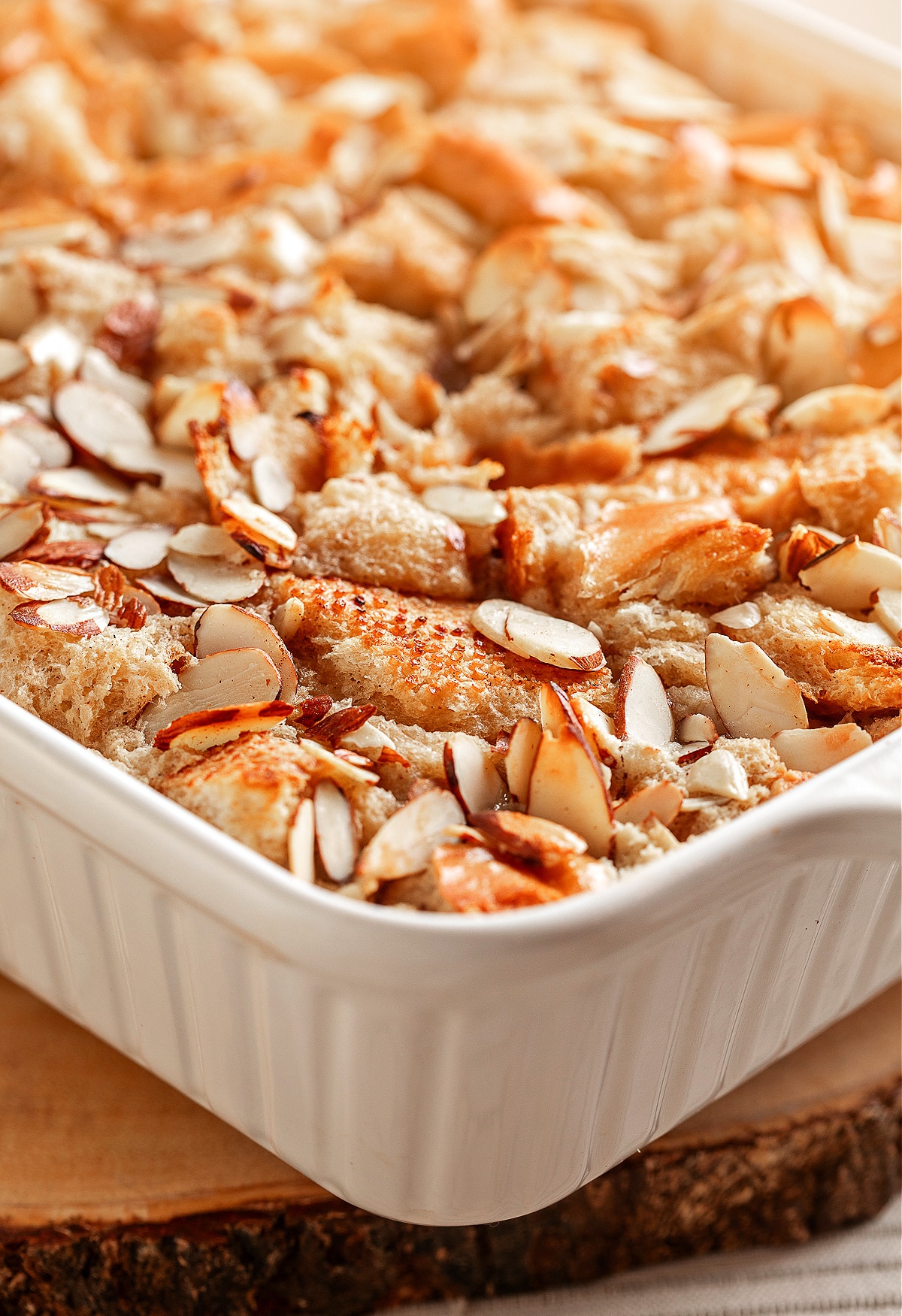 A close-up of almond cream bread pudding in a white baking dish, showing the toasted edges and scattered sliced almonds on top.