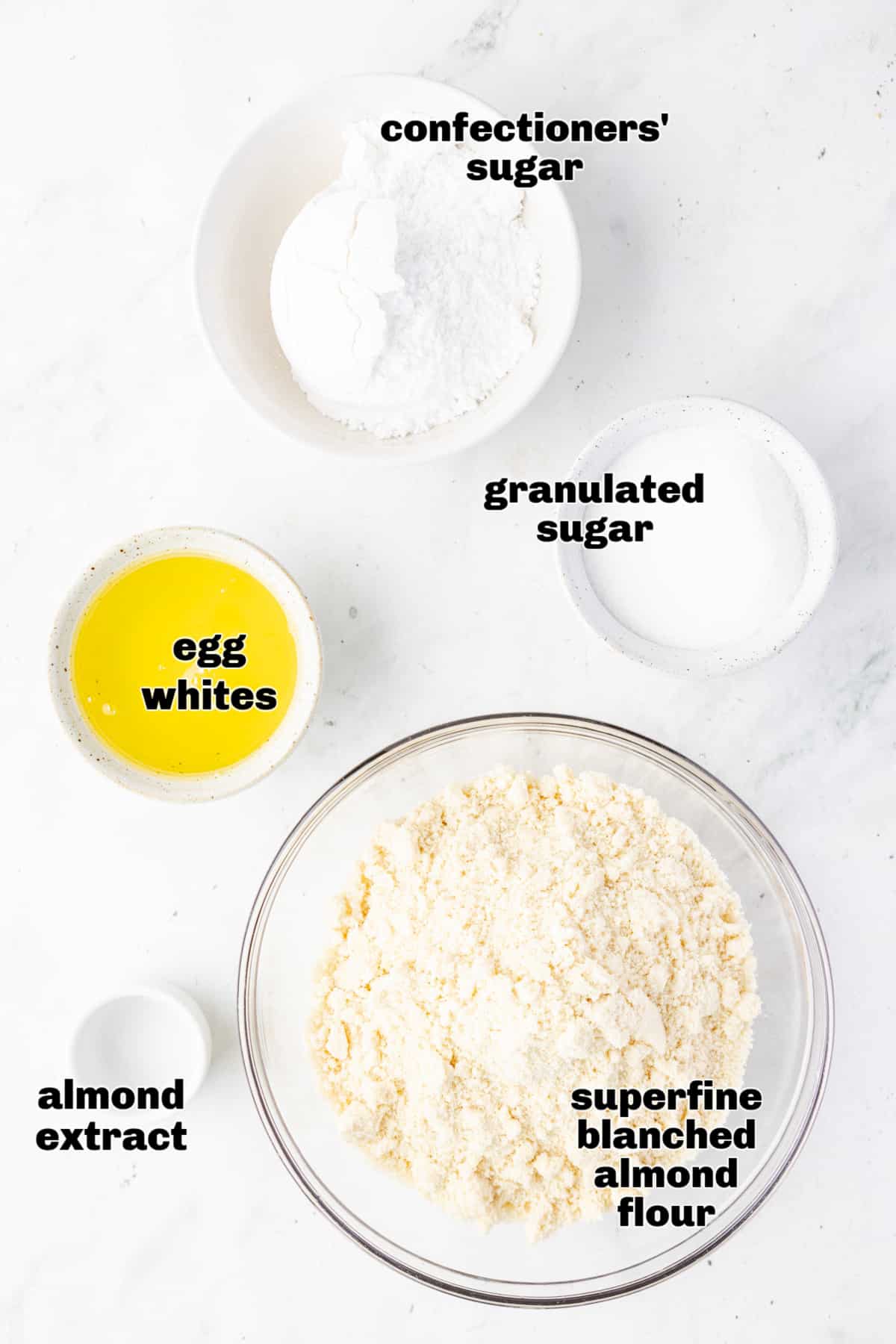 Ingredients for soft almond cookies arranged on a white surface, including almond flour, egg whites, sugars, and almond extract.