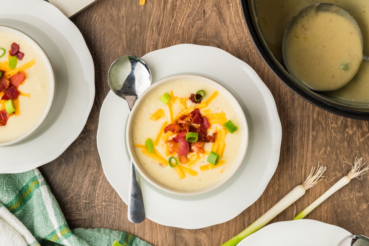 Creamy potato soup served with bacon, cheddar, and scallions ready for topping. Bowls of creamy potato soup topped with crisp bacon, shredded cheddar, and fresh scallions on a wooden table.