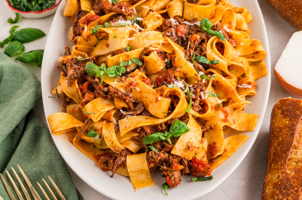 A large oval platter filled with tender beef ragù pappardelle, wide noodles twisted with shredded braised beef, tomato-rich sauce, fresh basil, and a sprinkling of parmesan.