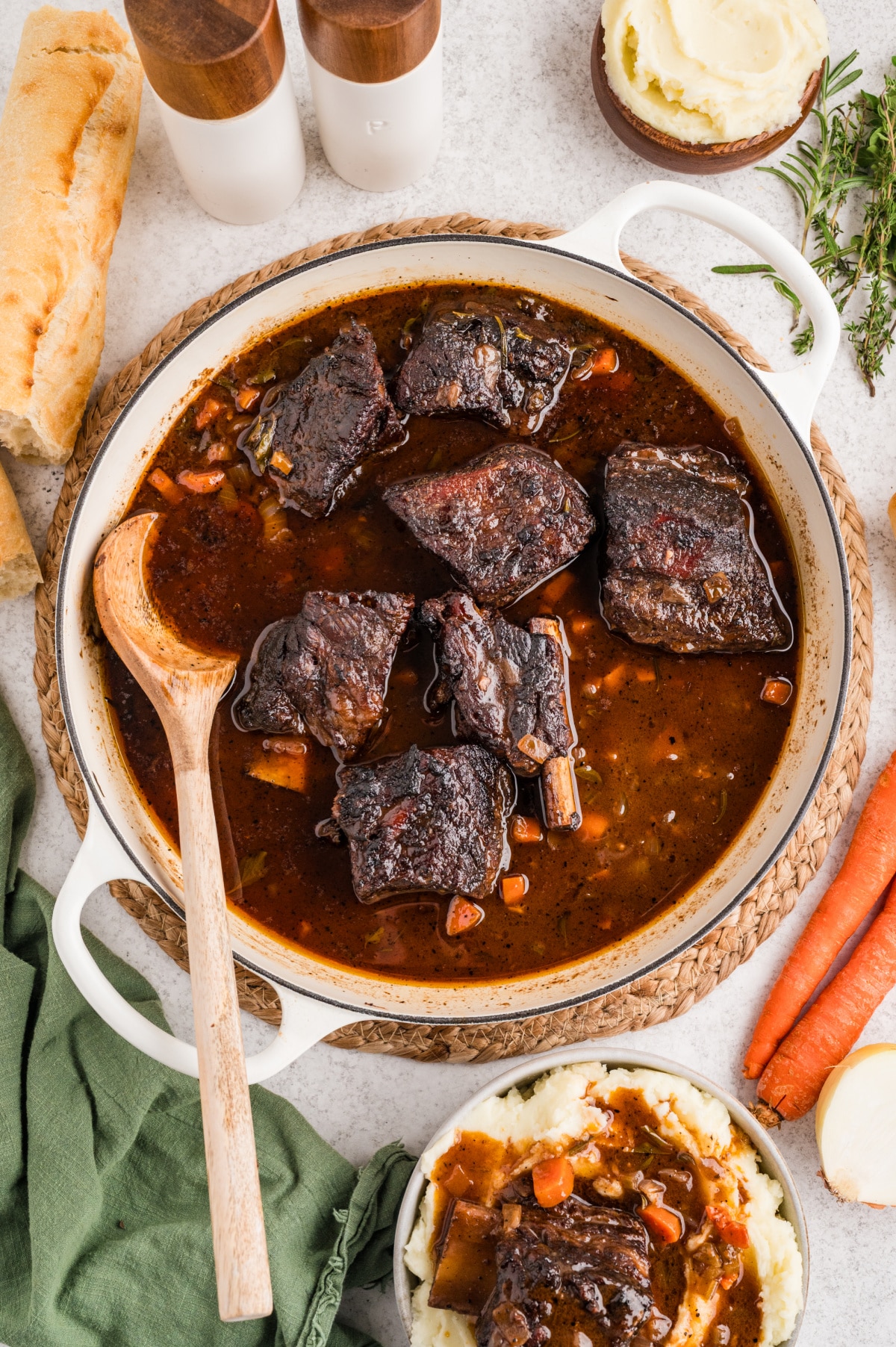 Braised short ribs resting in their cooking liquid with carrots and aromatics after slow oven cooking. Short ribs braising in a wide pan with a dark, glossy sauce, vegetables, and herbs after hours in the oven.