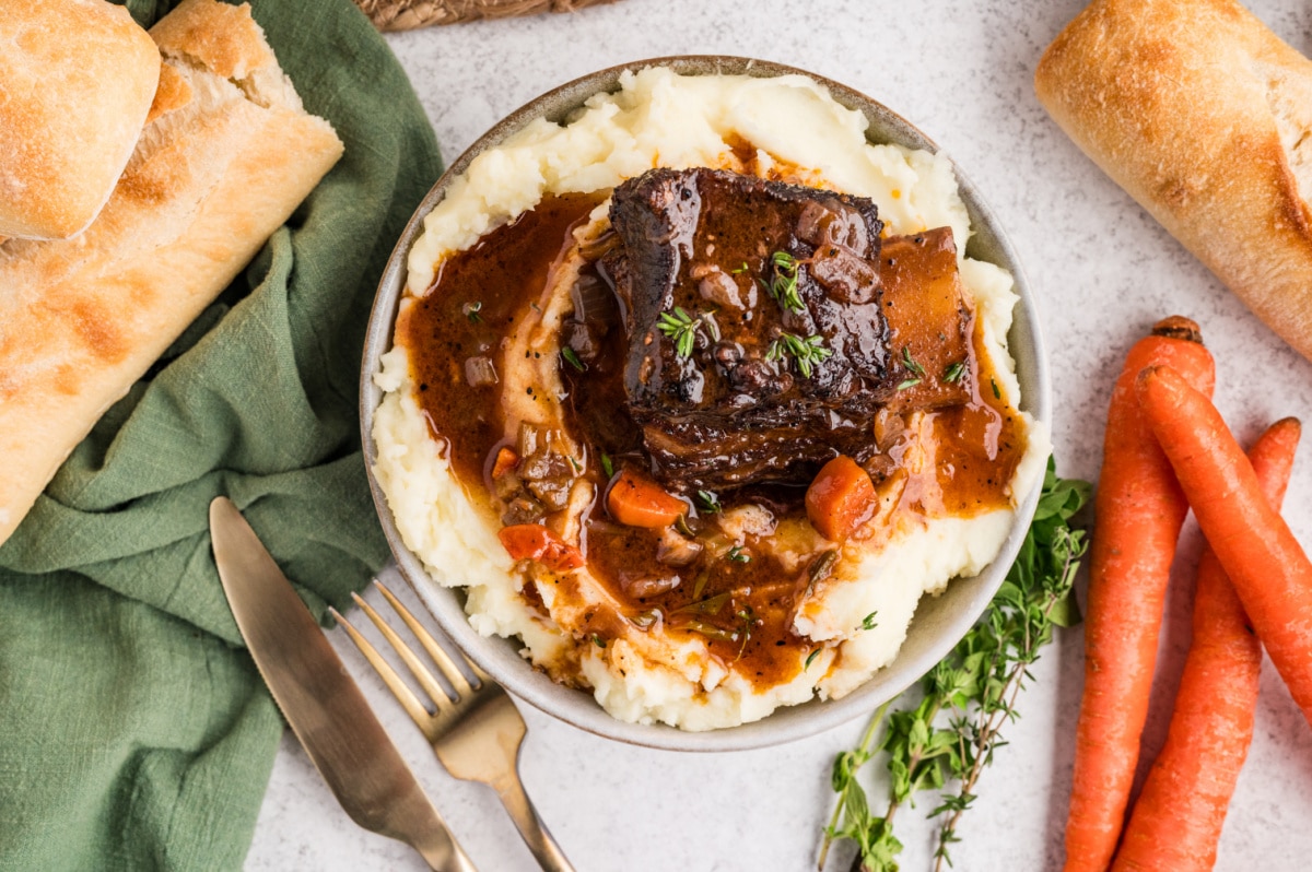 Braised beef short ribs plated over mashed potatoes with tender vegetables and rich pan sauce. Braised beef short ribs served in a bowl of mashed potatoes with carrots, herbs, and a deep, savory sauce.
