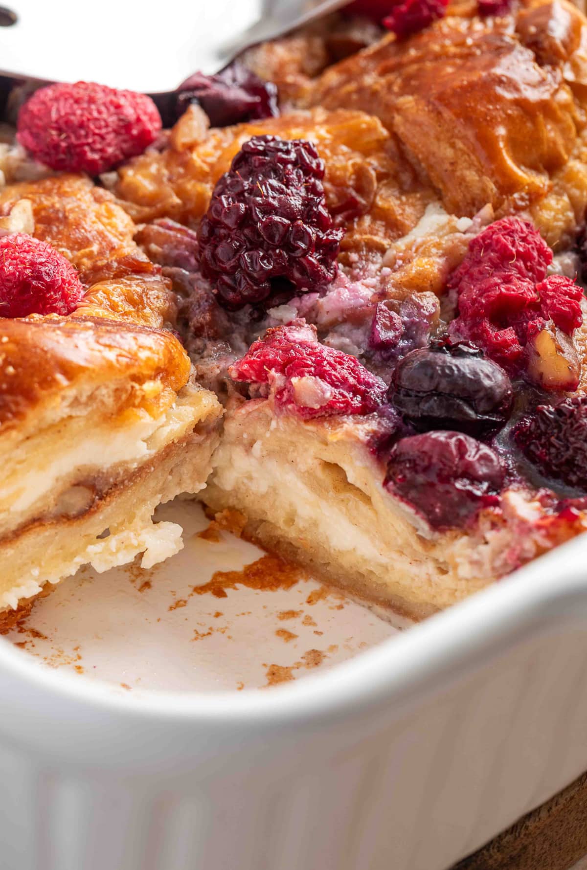 A close-up of baked croissants soaked in custard and topped with mixed berries and pecans inside a ceramic baking dish.