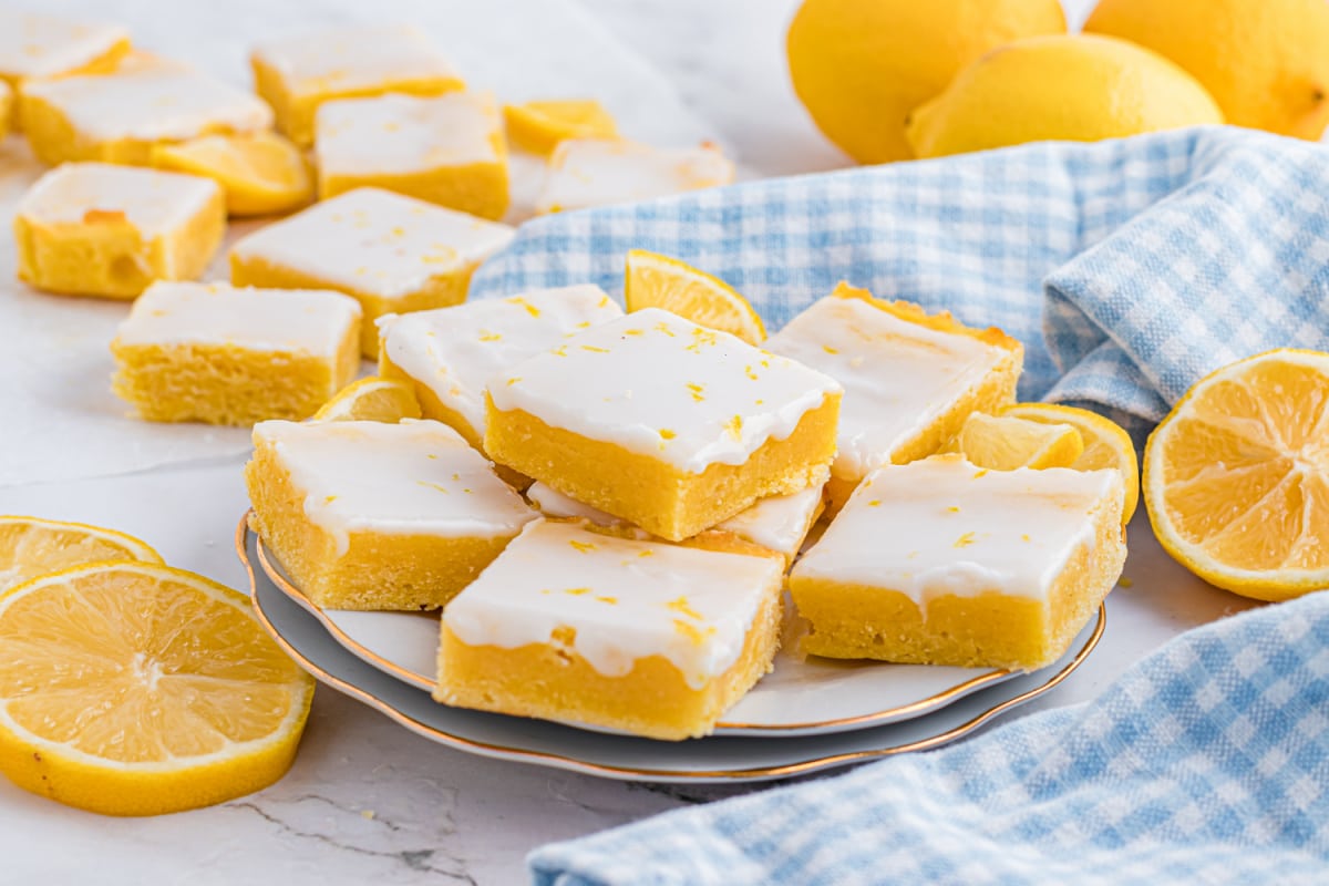 Stacked lemon dessert bars with a thick, buttery crumb and a glossy lemon glaze on top. Close-up of glazed citrus dessert bars stacked on a plate, showing a dense lemon crumb and smooth white glaze.