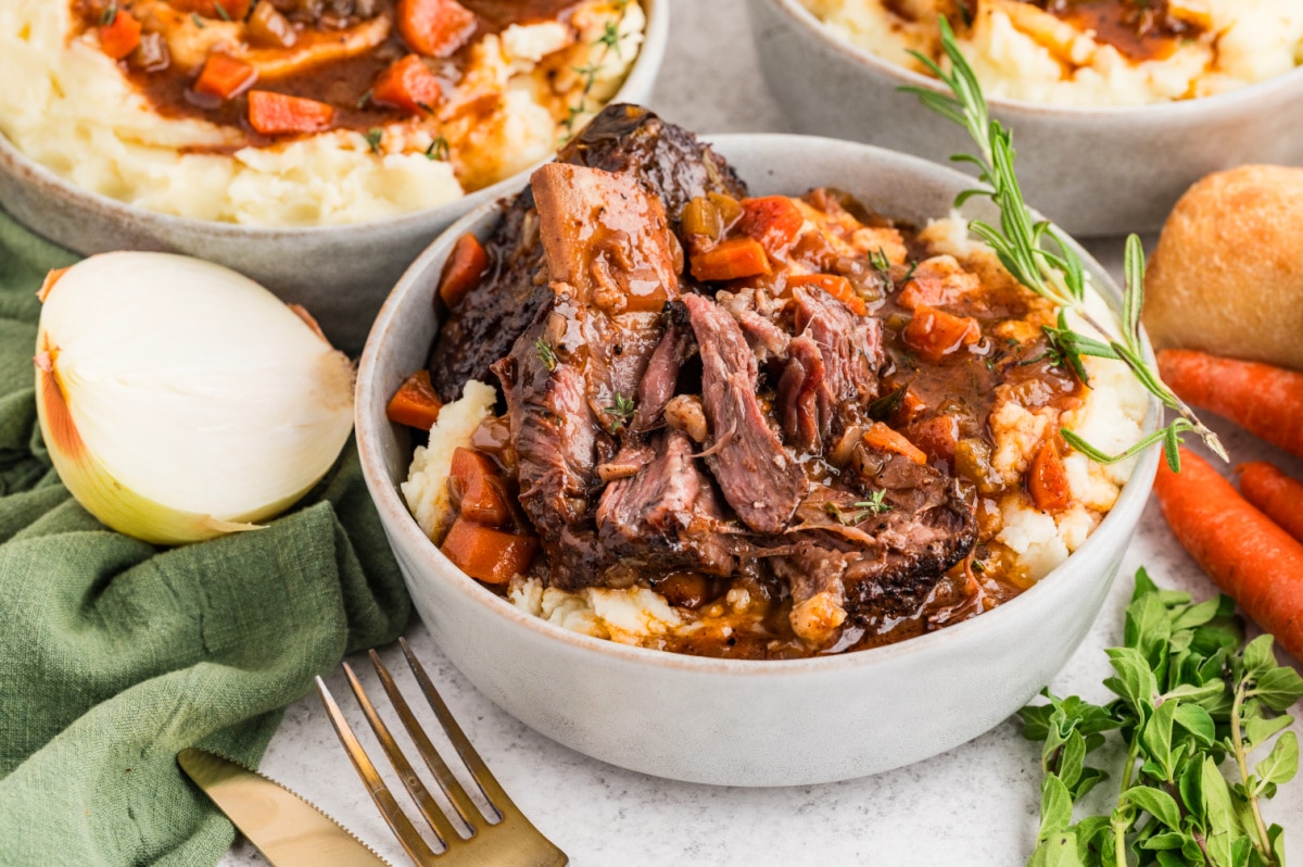 Braised short ribs shown fork-tender over mashed potatoes with vegetables and reduced cooking sauce. Tender braised short ribs pulled apart over mashed potatoes, surrounded by carrots, herbs, and thickened pan sauce.