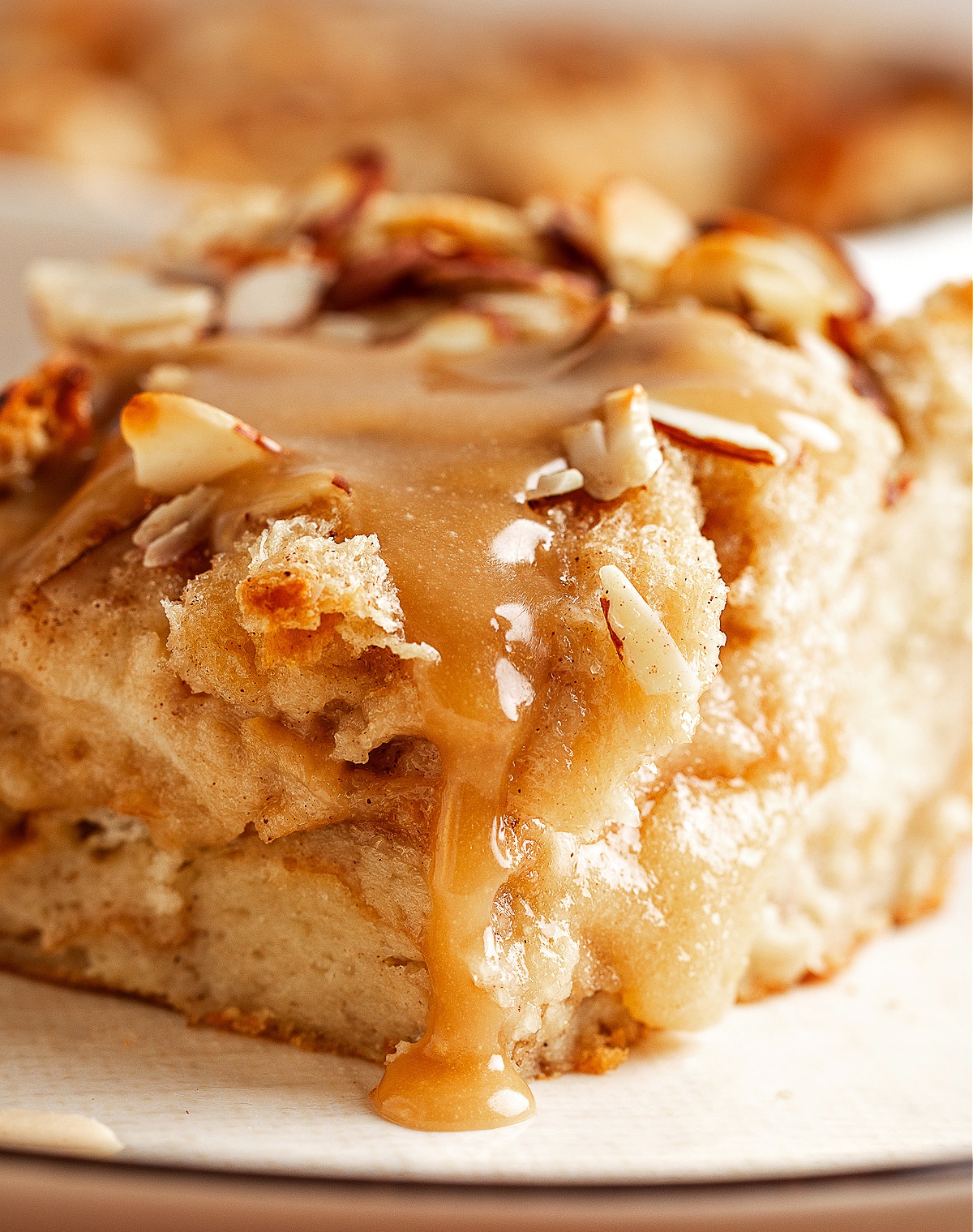 A close slice of almond bread pudding with warm brown sugar glaze dripping down the side, showing the soft custard interior and toasted almond topping.