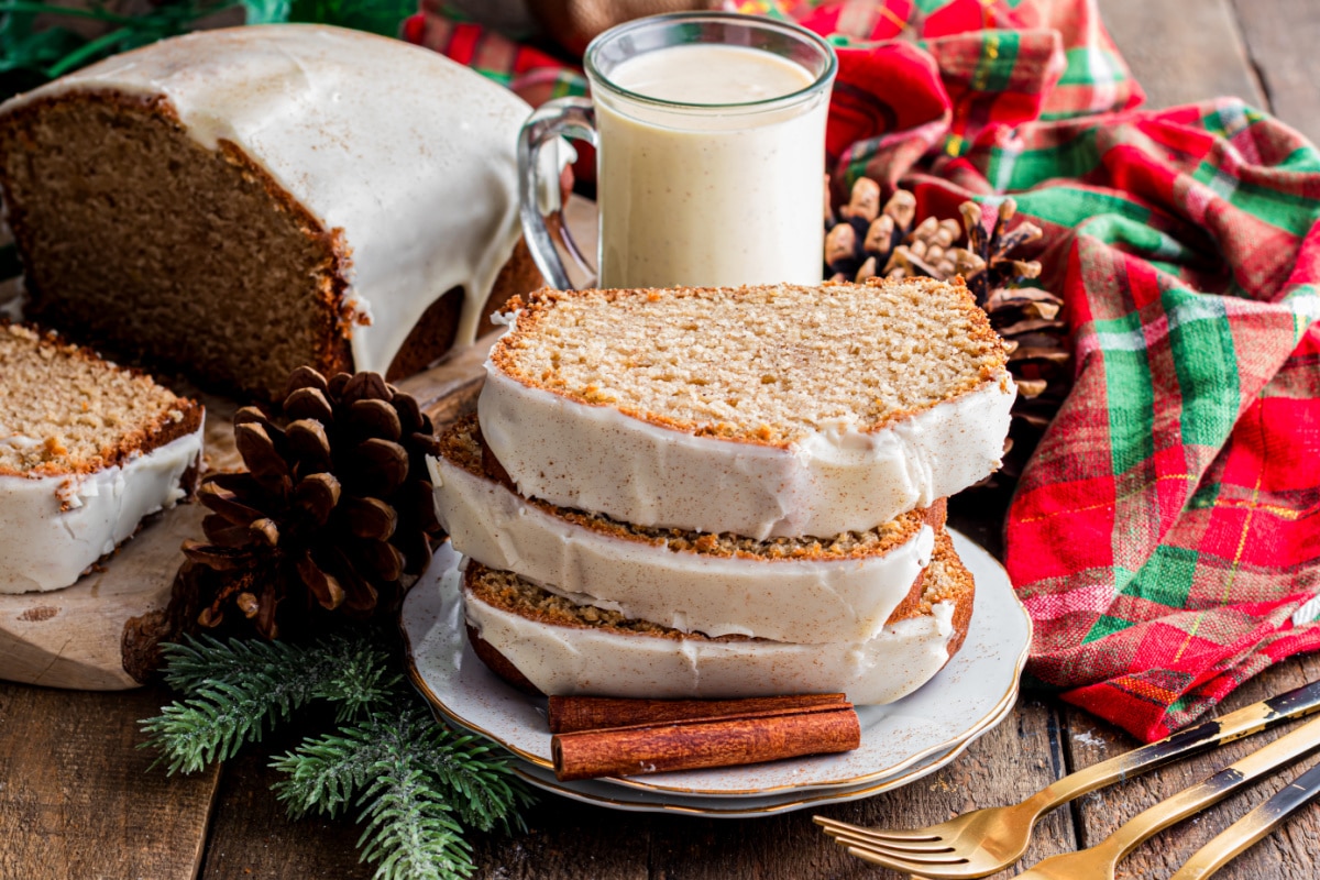 Eggnog bread slices with glaze and warm holiday spices Sliced eggnog bread stacked on a plate with a set glaze, surrounded by pinecones, cinnamon sticks, and a mug of eggnog on a holiday table.