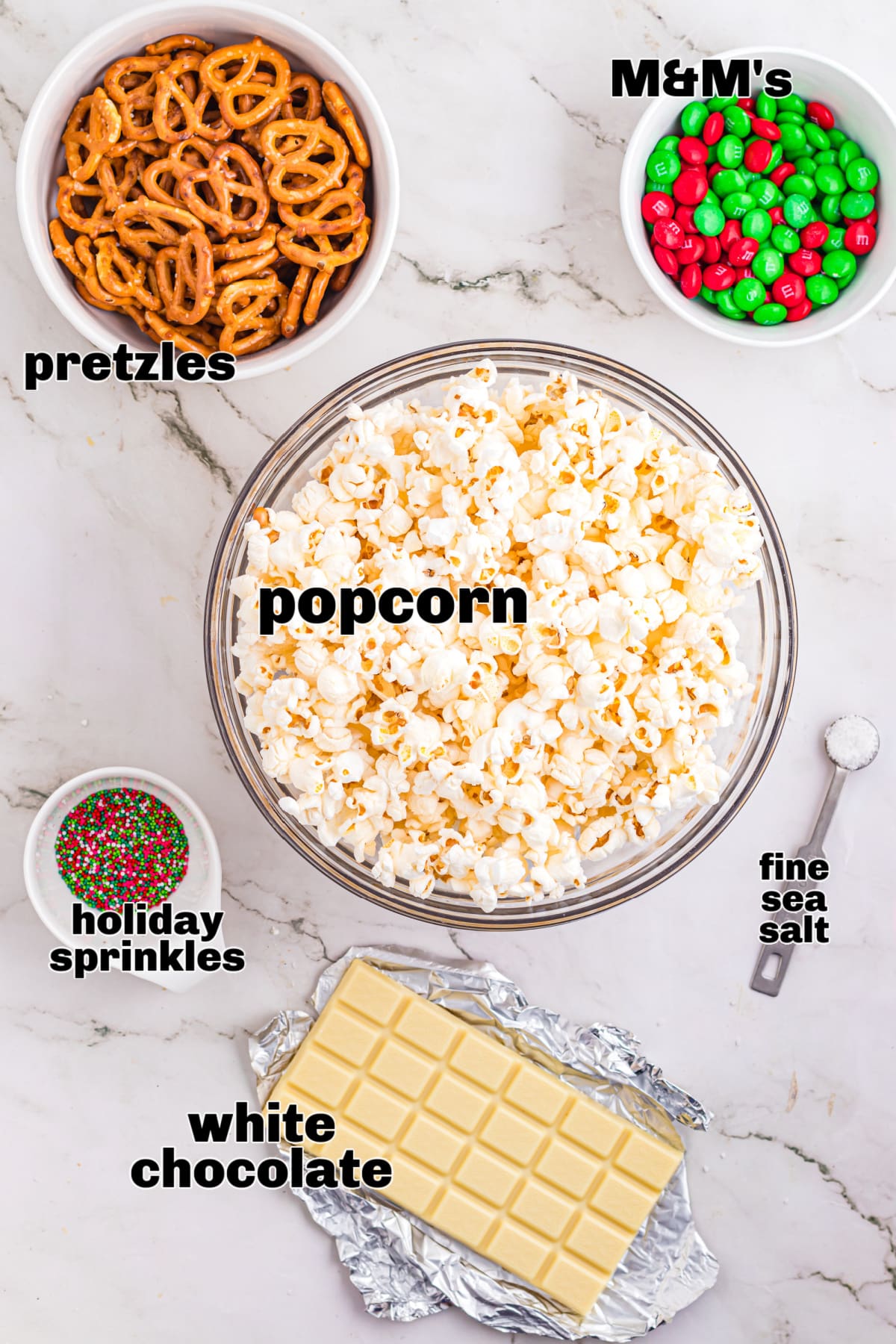 The ingredients for Christmas popcorn mix are arranged on a light countertop. Ingredients for Christmas popcorn mix including plain popcorn, mini pretzels, white chocolate, holiday sprinkles, and candy-coated chocolates.