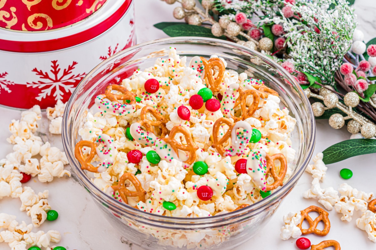 A glass bowl filled with holiday popcorn mixed with white chocolate pretzels and candy. Holiday popcorn mix with white chocolate–dipped pretzels, red and green candy-coated chocolates, and sprinkles in a glass bowl.
