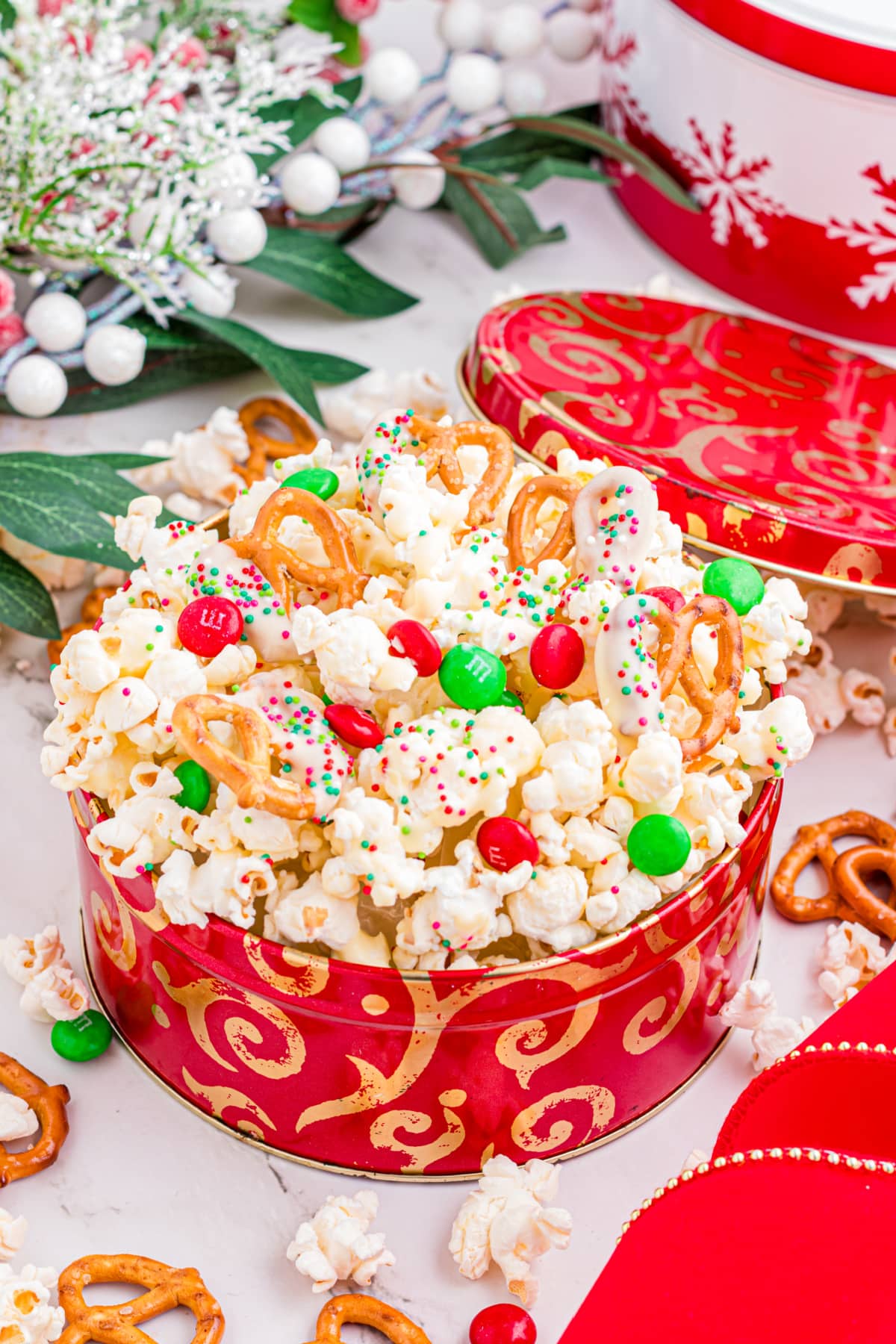 This white chocolate holiday popcorn is packed into a red tin with pretzels and candy. White chocolate popcorn with pretzels, holiday M&M’s, and sprinkles piled into a red Christmas tin