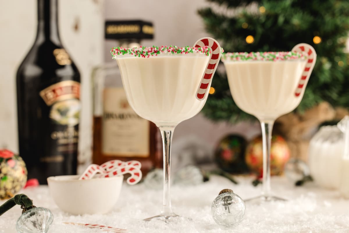 Two sprinkle-rimmed holiday martinis with candy cane stirrers in a festive winter scene. Two creamy holiday martinis with sprinkle rims and candy cane stirrers set on a snowy tabletop, with liqueur bottles and a Christmas tree softly blurred in the background.