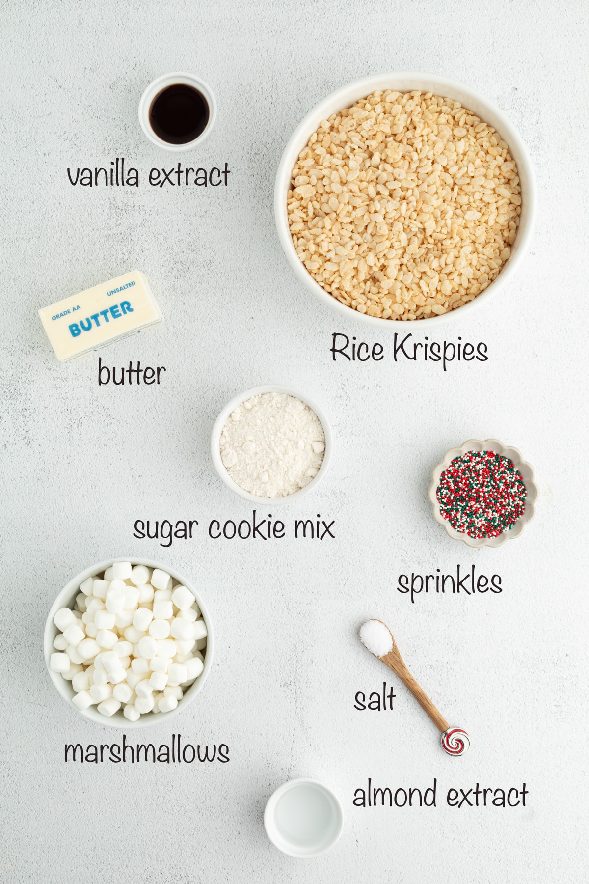 Ingredients for sugar cookie–flavored Rice Krispie treats arranged on a light surface, including marshmallows, cereal, butter, sugar cookie mix, extracts, salt, and holiday sprinkles.