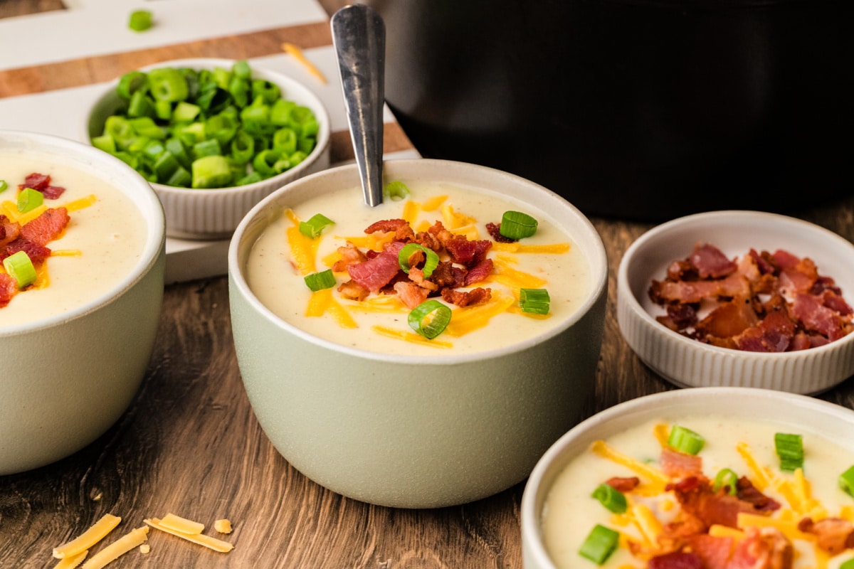 Bacon cheddar potato soup served warm with scallions and a ladle nearby. A bowl of bacon cheddar potato soup garnished with scallions, set on a plate beside the pot and ladle.