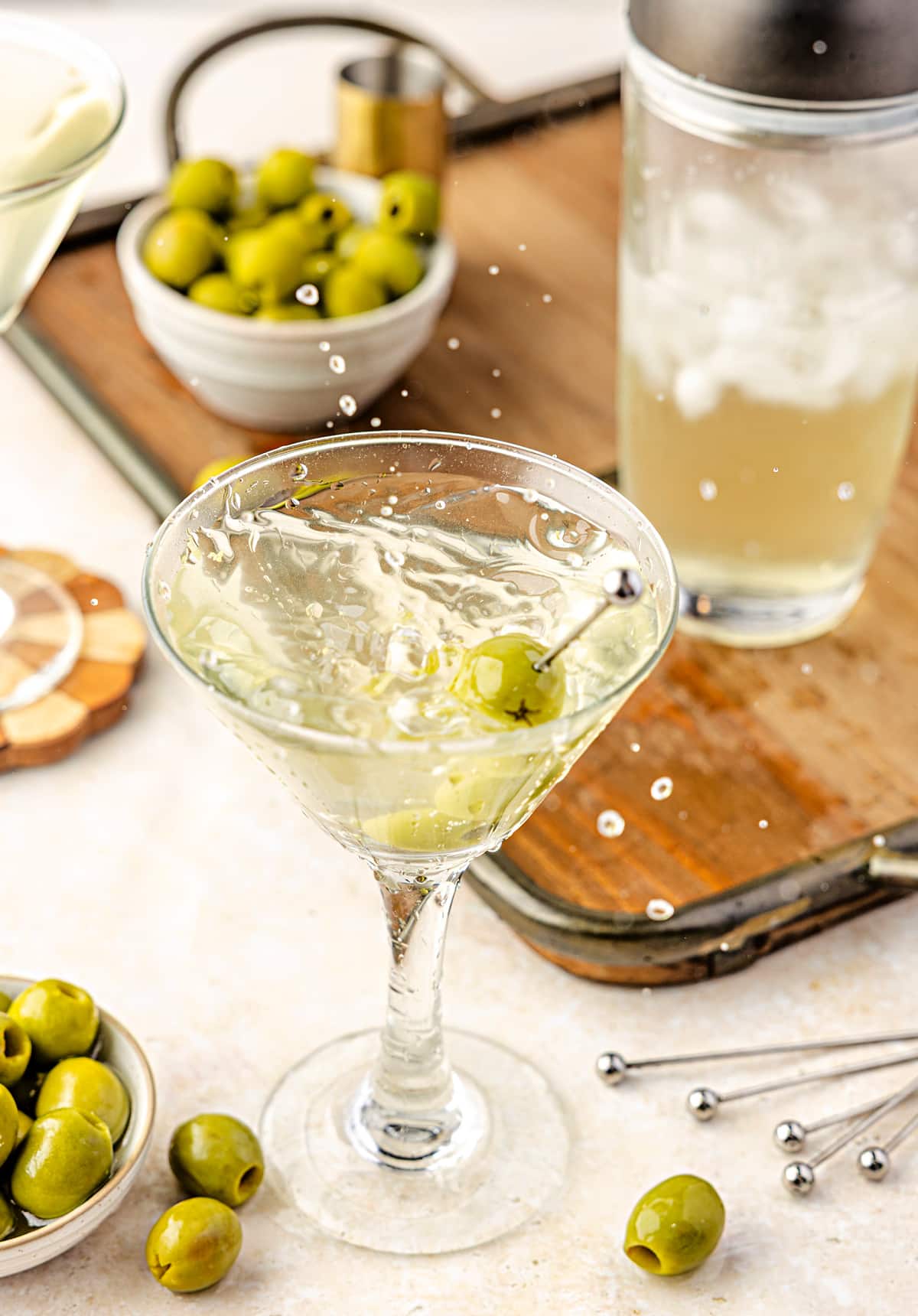 An extra dirty martini splashing into a chilled glass, finished with green olives and surrounded by olive brine, ice, and bar tools.
