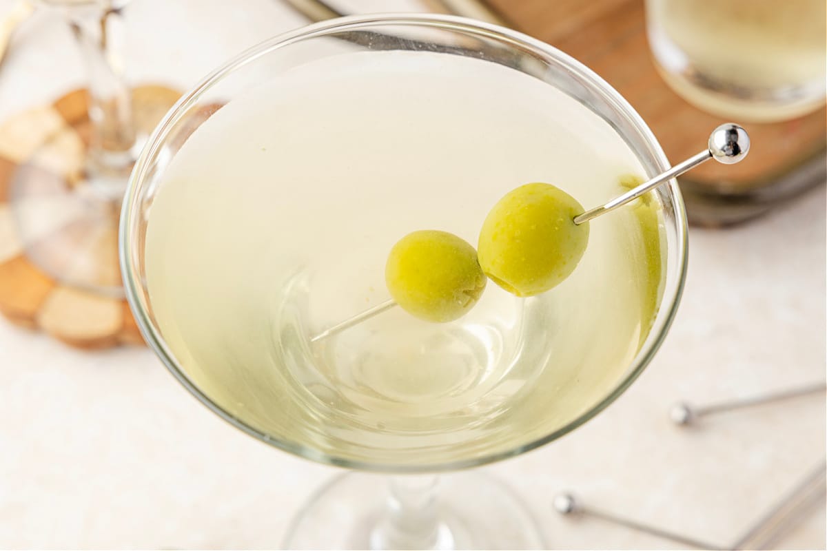 A chilled martini glass filled with a pale, briny cocktail and green olives, set against a shaker and bowls of olives in the background.