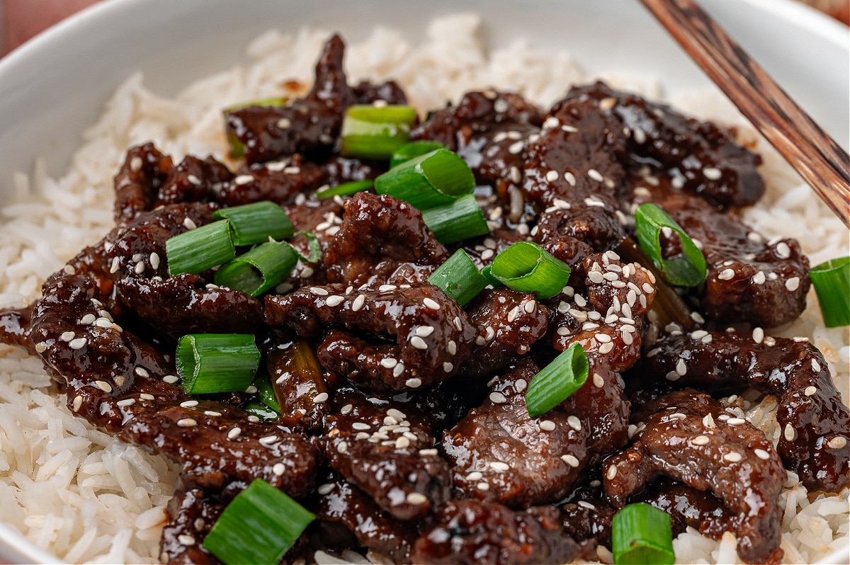 Close up of glossy sliced beef in a ginger soy sauce spooned over rice and finished with sesame seeds.