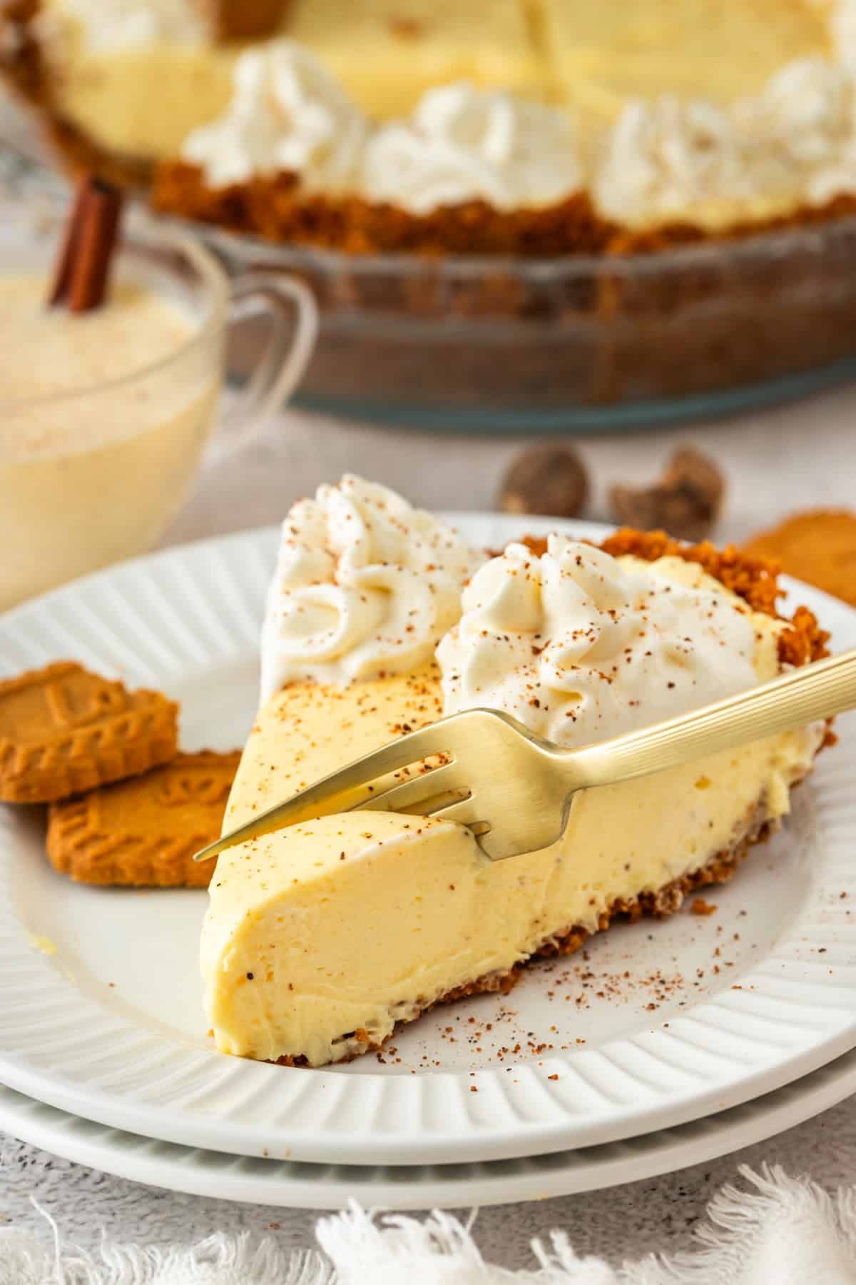A plated slice of eggnog cream pie topped with whipped cream and nutmeg, served with a fork and cookies on the side.