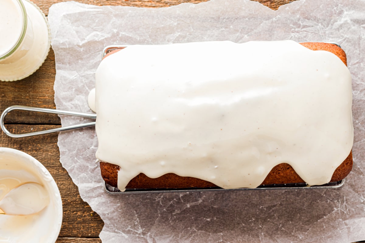 Eggnog quick bread cooling with smooth eggnog glaze Freshly glazed eggnog quick bread resting on a cooling rack, with a smooth white glaze cascading over the top on parchment paper.