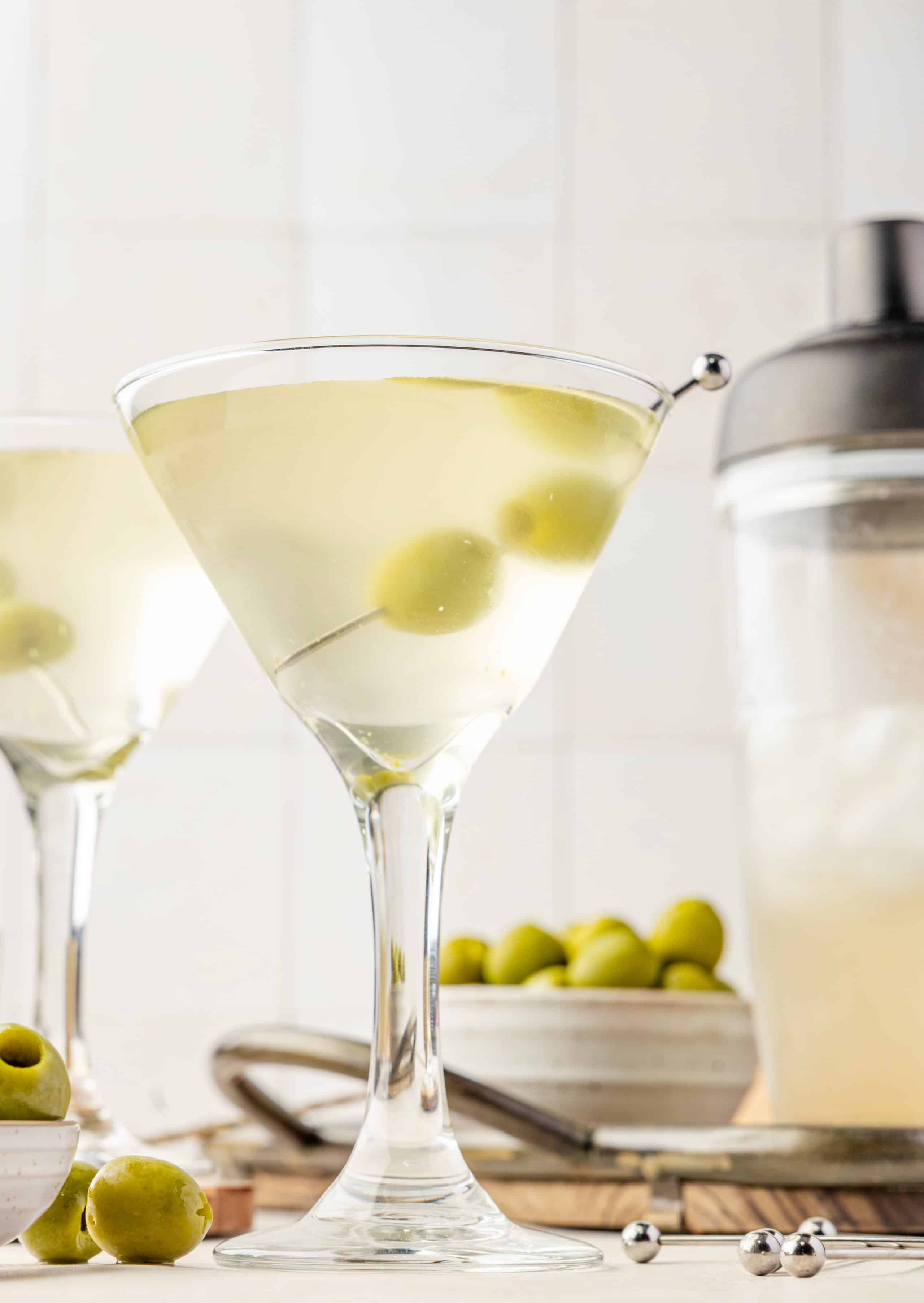 An extra dirty martini resting on a wooden tray, garnished with two green olives and surrounded by olive brine, ice, and cocktail picks.