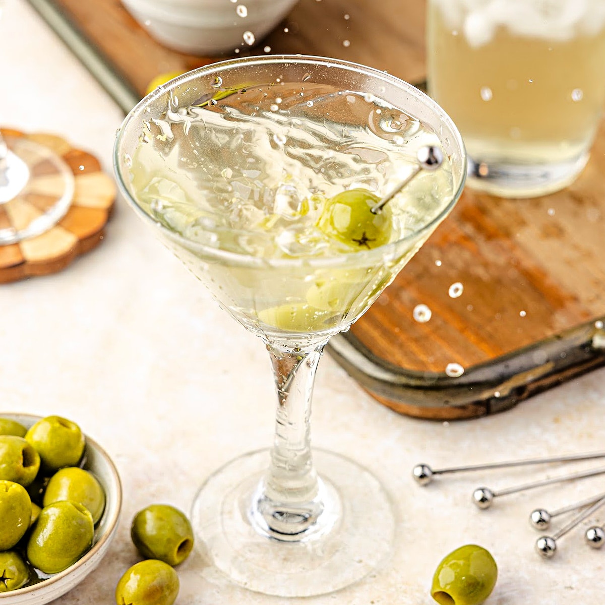 Extra dirty martini splashing into a chilled glass with green olives, olive brine, and bar tools on the counter.