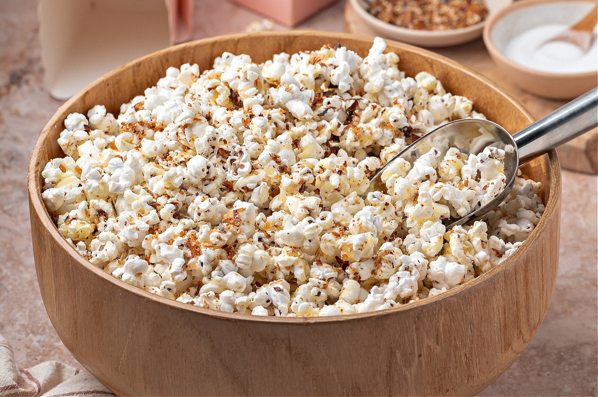 A wooden bowl filled with popcorn coated in a sweet and spicy seasoning blend, with a metal scoop resting inside.