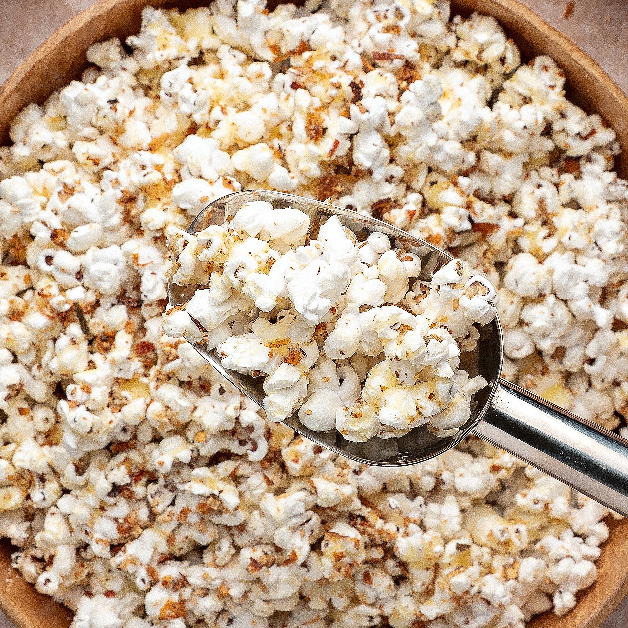 A metal scoop resting in a wooden bowl filled with sweet and spicy seasoned popcorn dotted with furikake flakes.