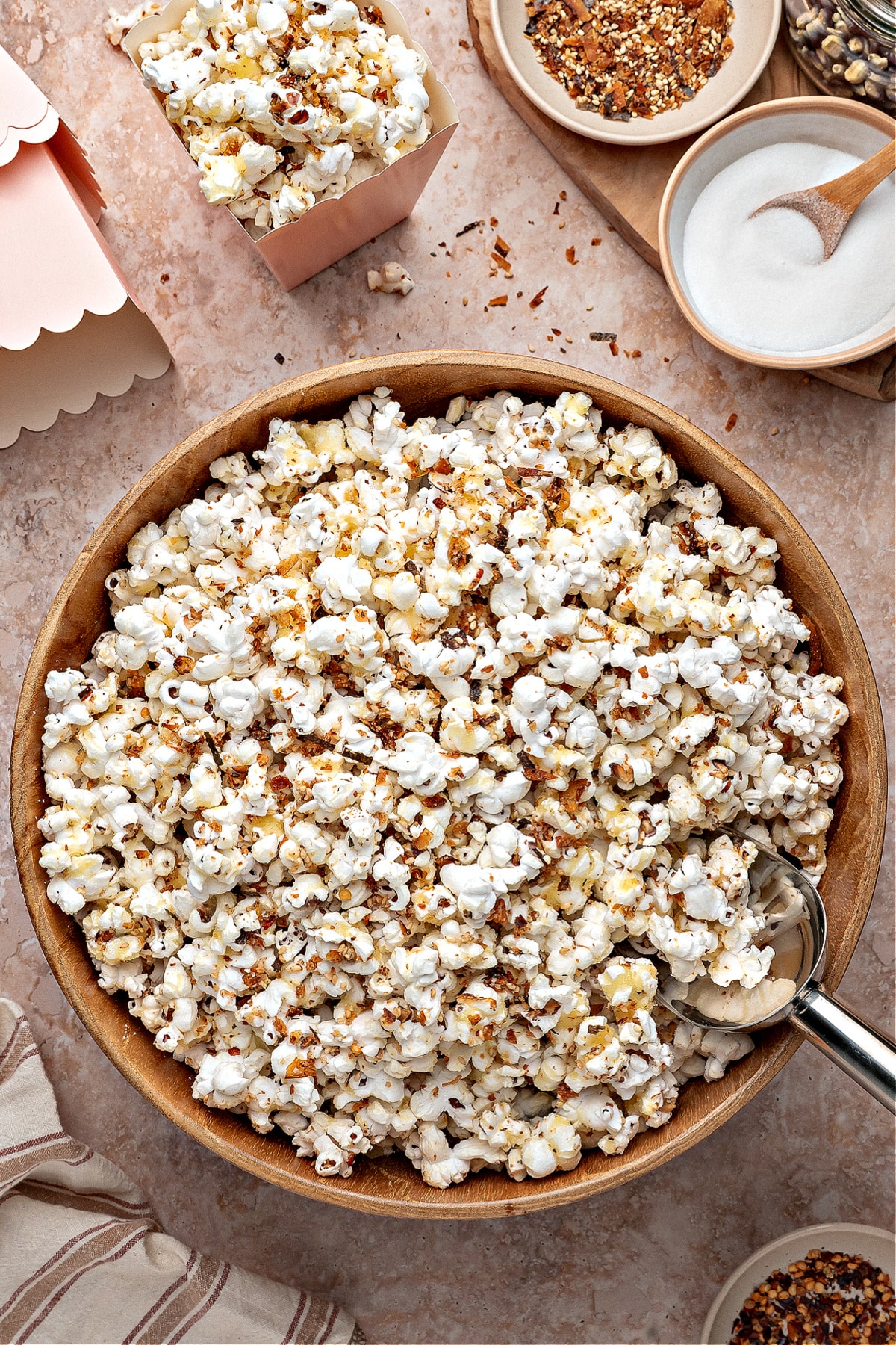 A large wooden bowl of sweet and spicy furikake popcorn sits on a stone surface, with scattered seasoning, small serving containers, and ingredients visible around it.