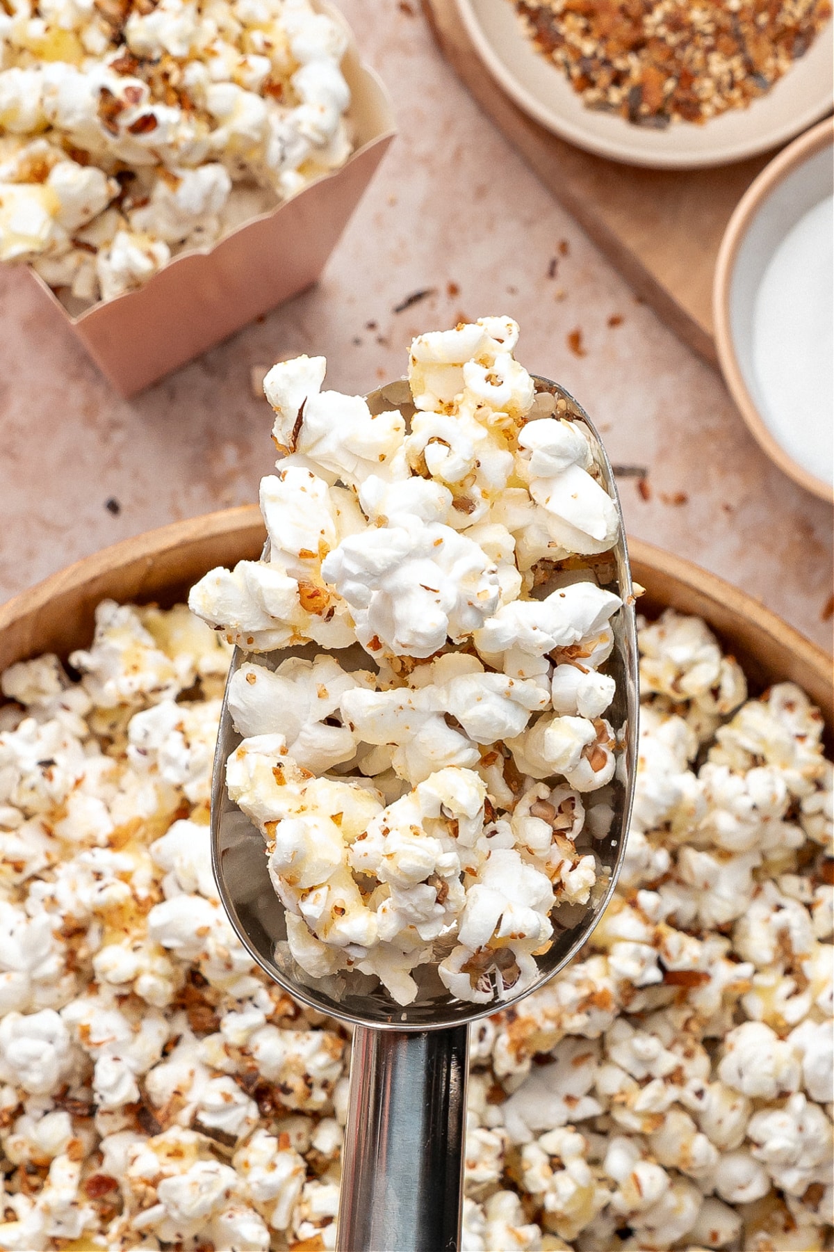 A metal scoop lifting a serving of seasoned popcorn above a wooden bowl, with toppings and ingredients in the background.